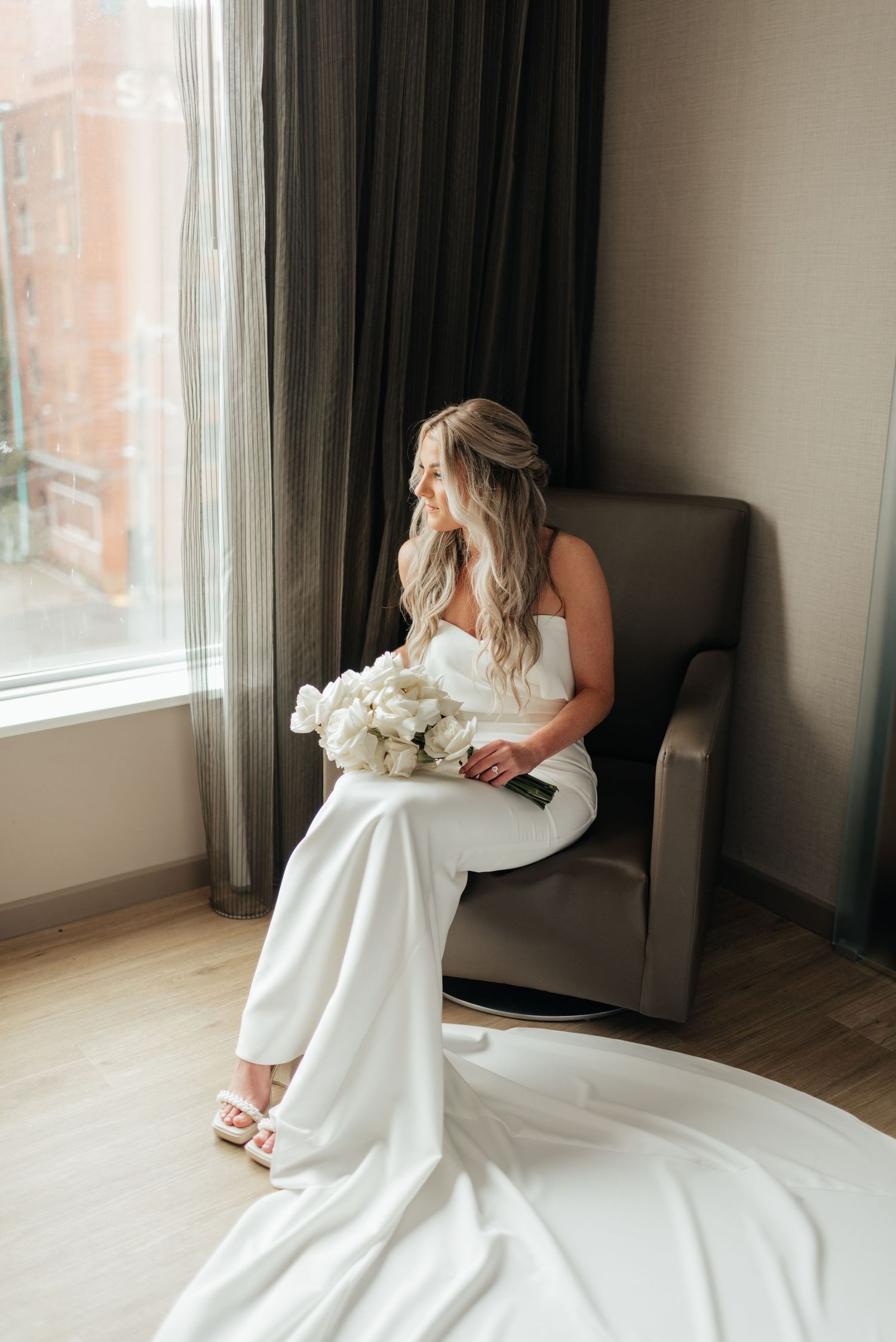 A bride in a flowing white wedding gown twirls and dances in front of large windows in a minimalist room.