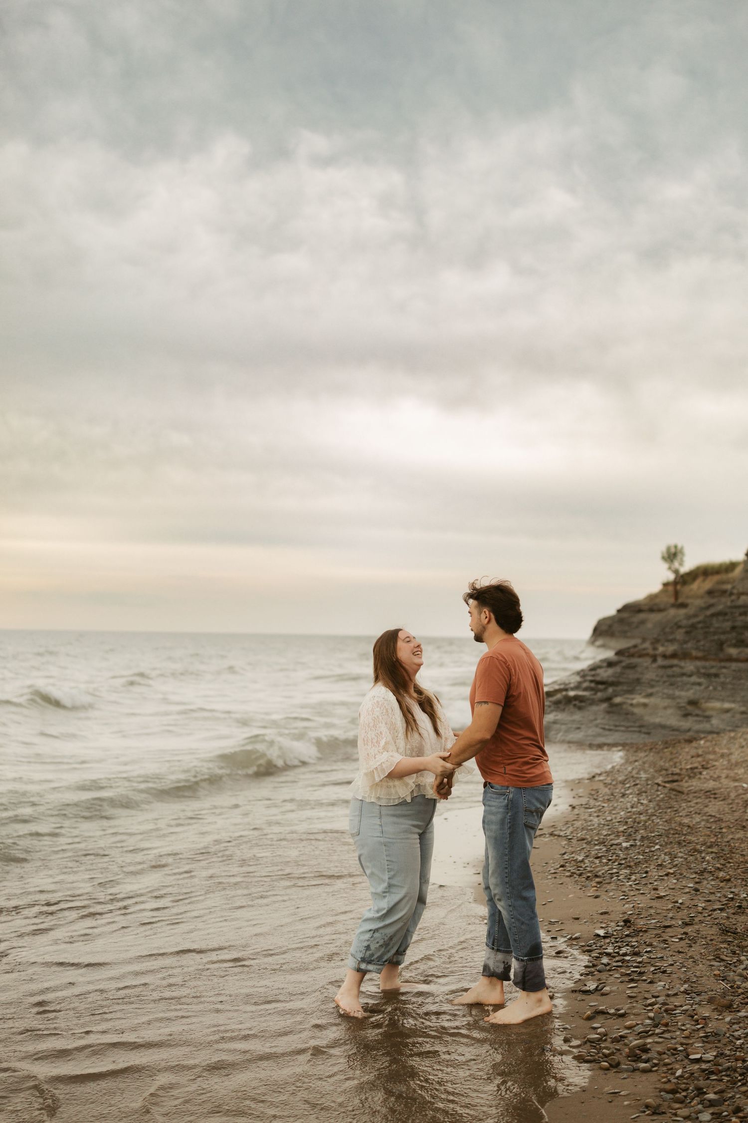 Couple holding hands barefoot on a rocky beach shore at sunset with cloudy skies and ocean waves.