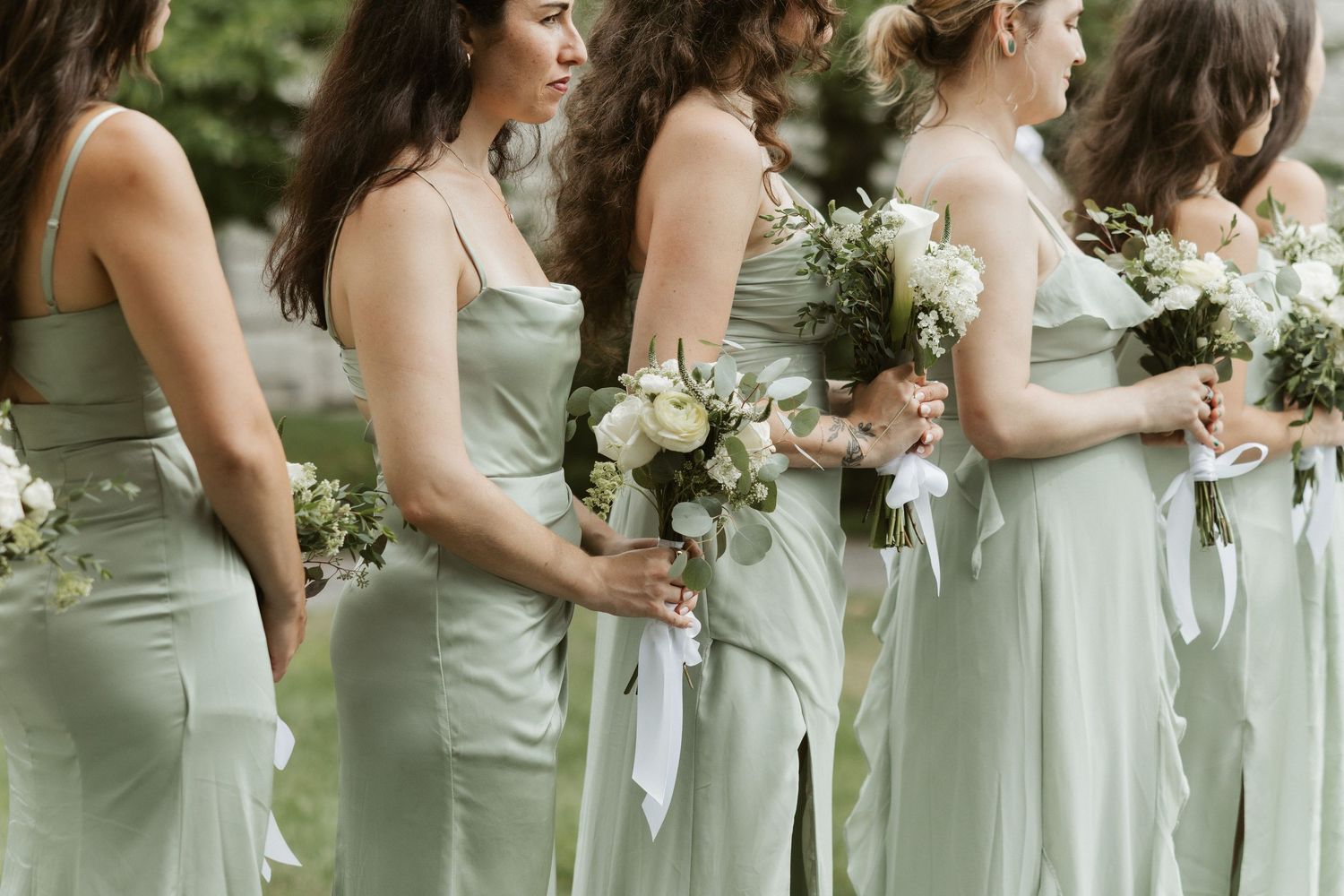 Row of bridesmaids in sage green dresses holding white bouquets during outdoor wedding ceremony.