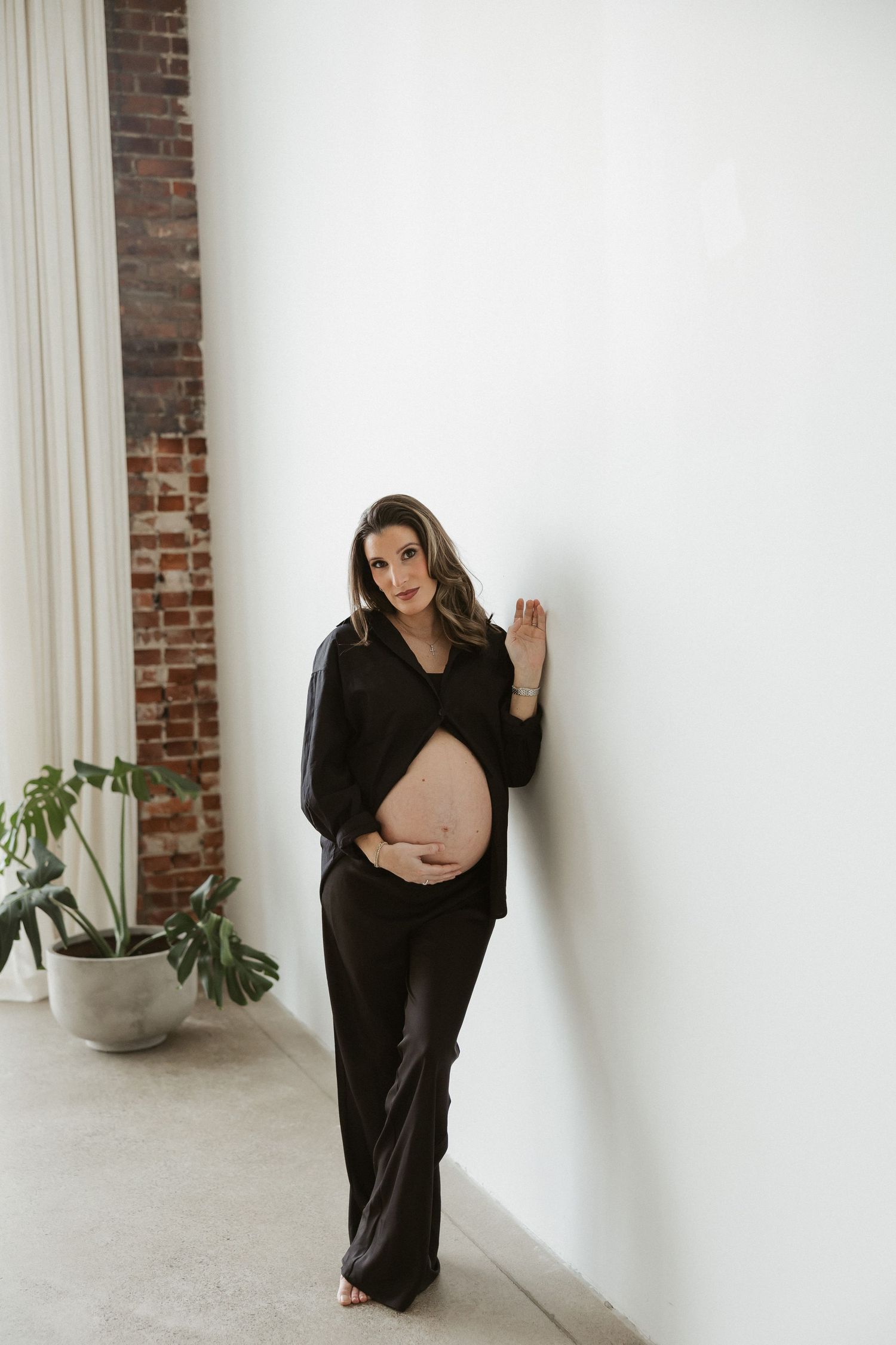 Pregnant woman in black outfit leans against white wall cradling baby bump in bright studio with brick wall.