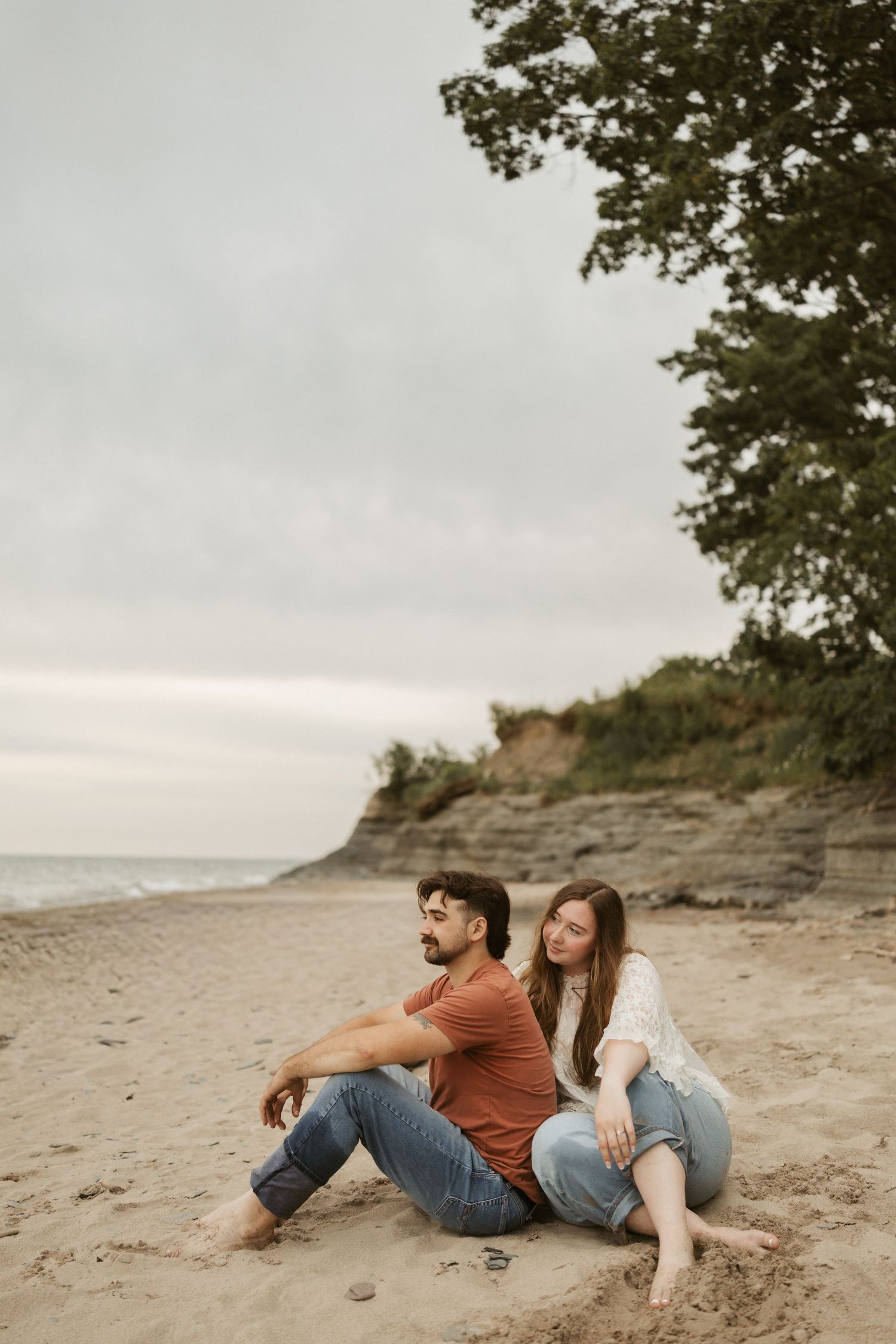 Couple sitting together on sandy beach near cliffs, woman leaning on man's back, casual jeans outfits, cloudy sky.