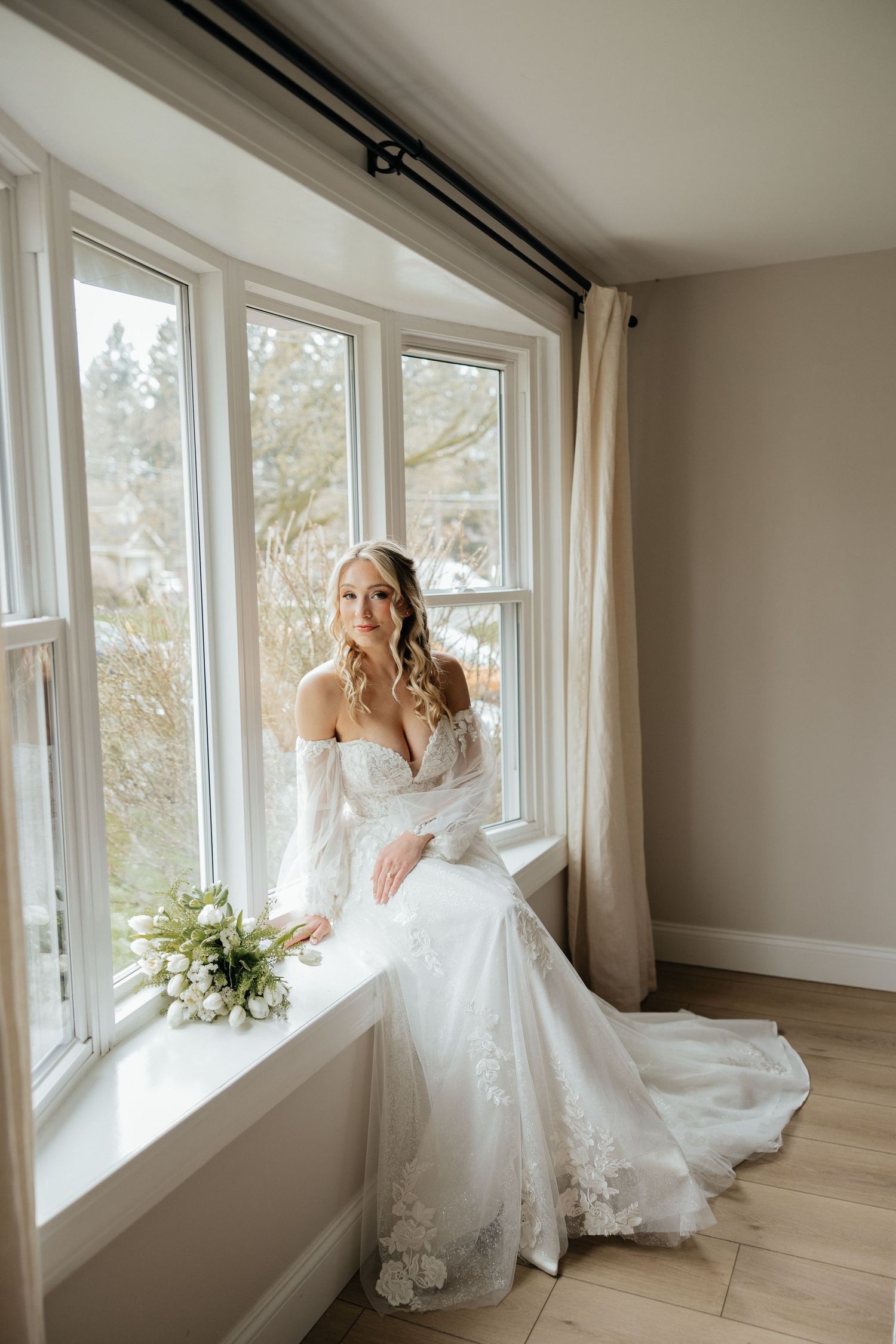 Elegant bride in flowing white wedding dress poses gracefully by bright sunlit windows in modern interior.