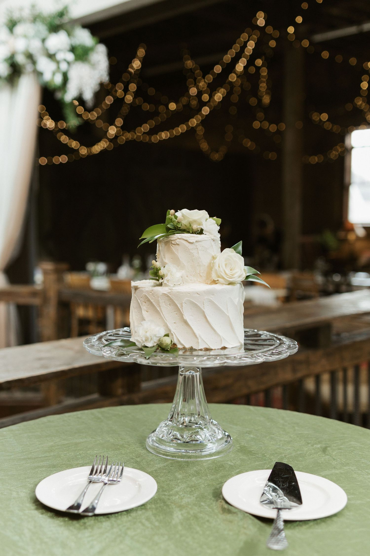 Two-tier white textured wedding cake with floral decorations displayed on crystal cake stand with string lights in background.