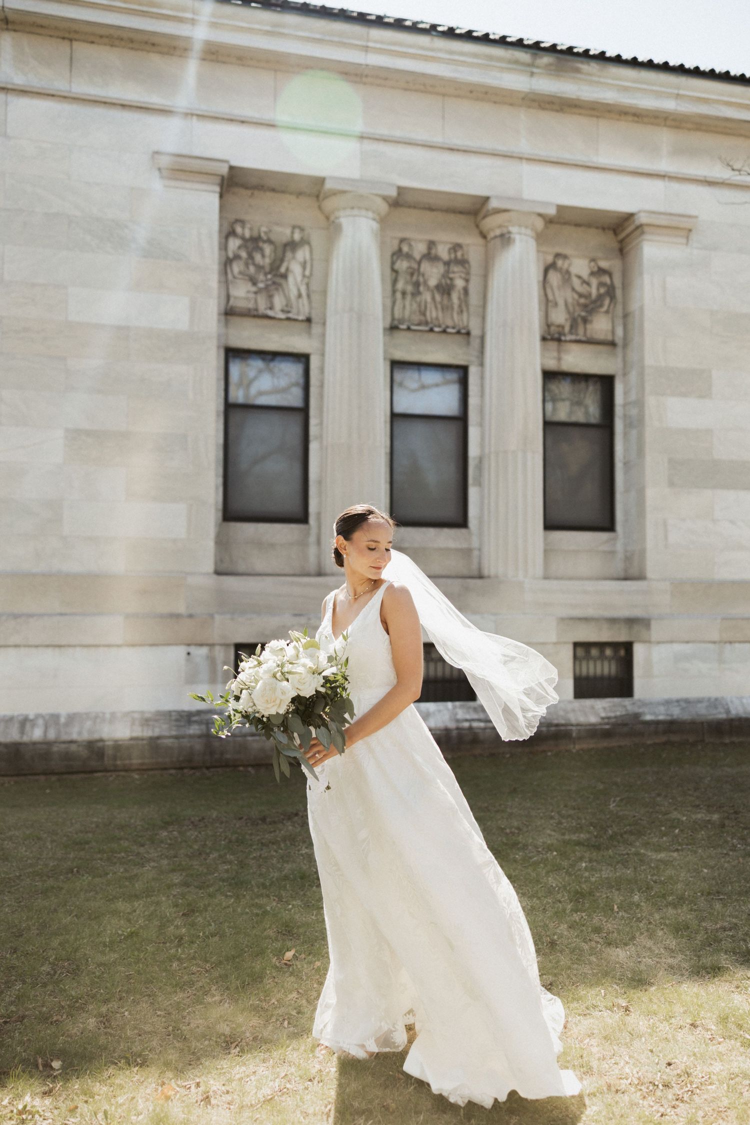 Bride in elegant white gown with veil flowing in wind holds white bouquet outside neoclassical building.
