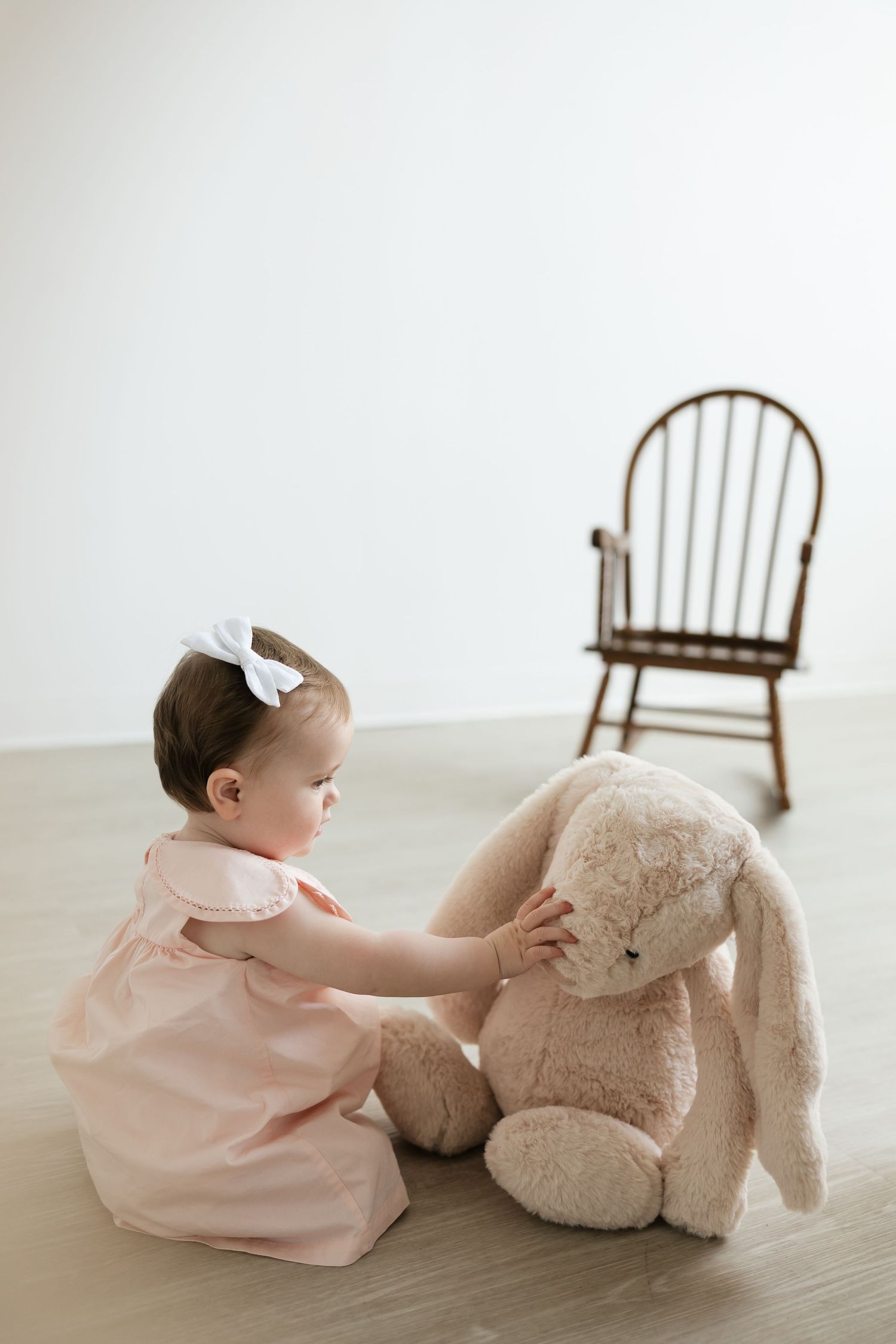 A baby girl in a pink dress with a white bow sits on the floor playing with a large stuffed bunny toy.