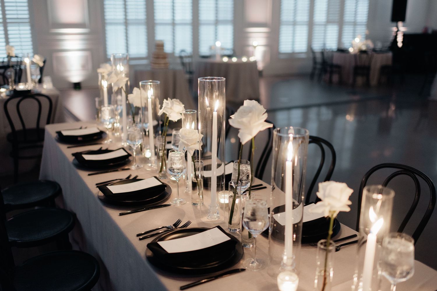 Elegant wedding reception table with tall glass candle holders, black chairs, and white place settings in modern venue.