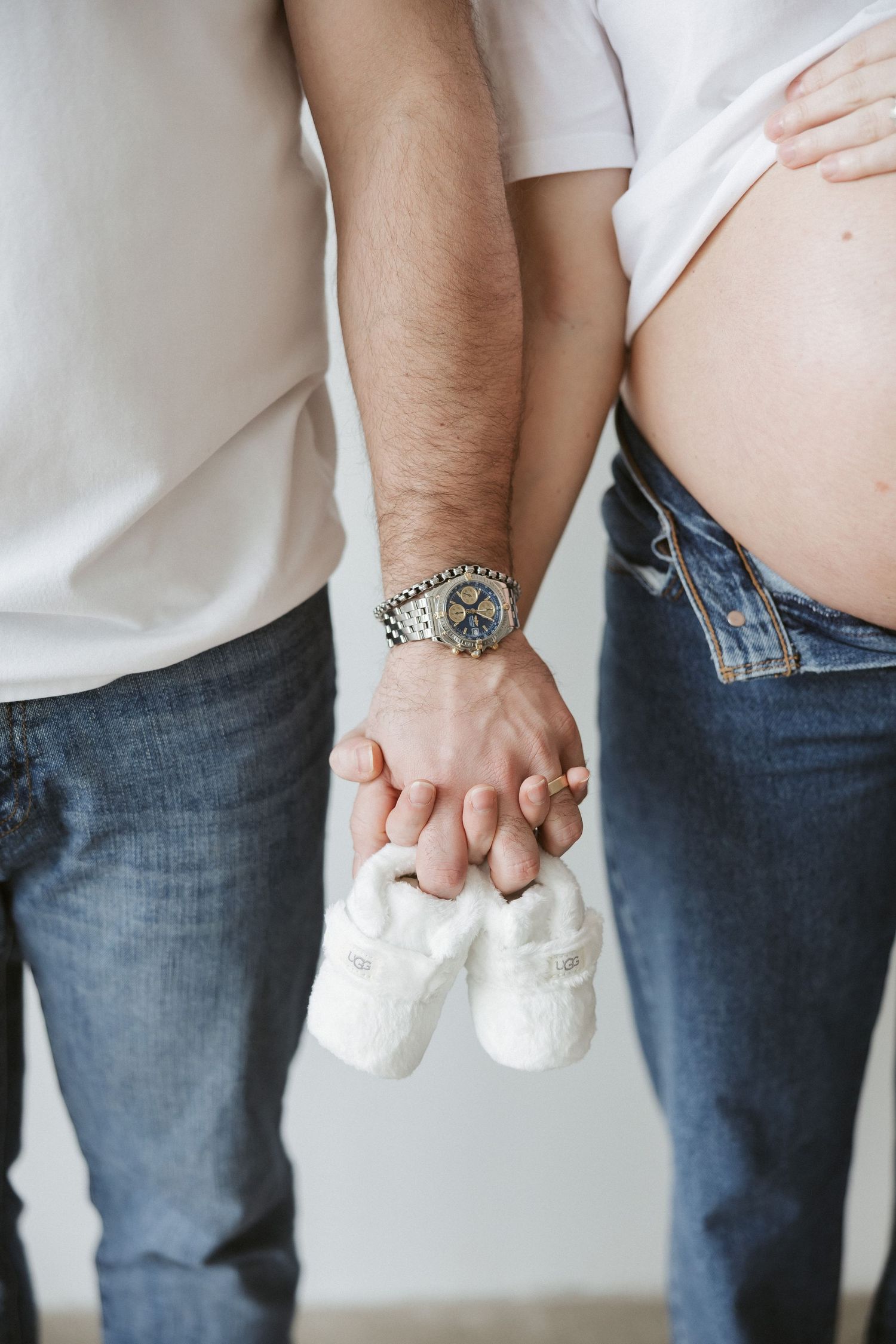 Expecting couple in jeans holds tiny white baby booties together, showing pregnant belly in close-up shot.