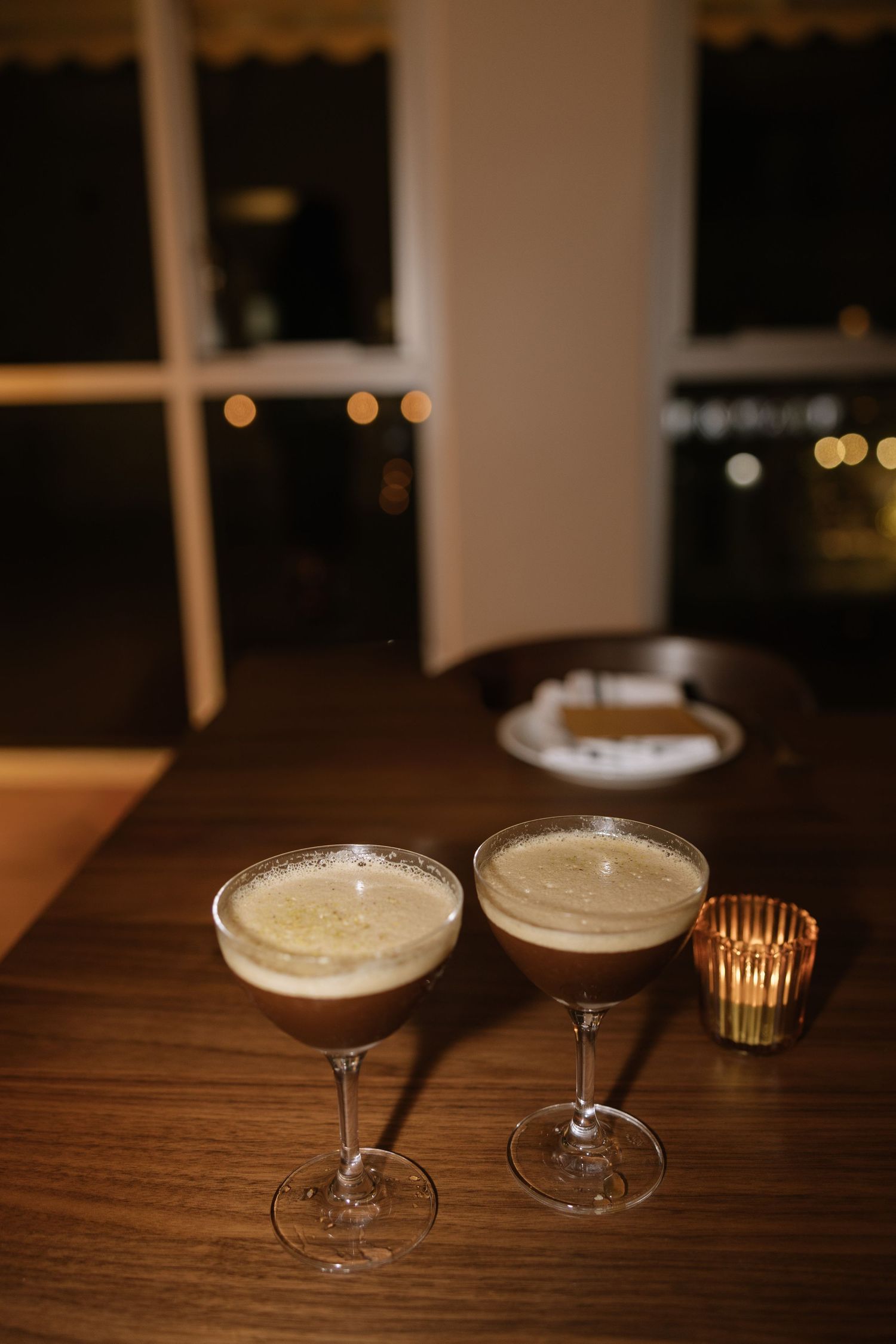 Two espresso martini cocktails with frothy tops on a wooden bar table, with bokeh lights in the background.