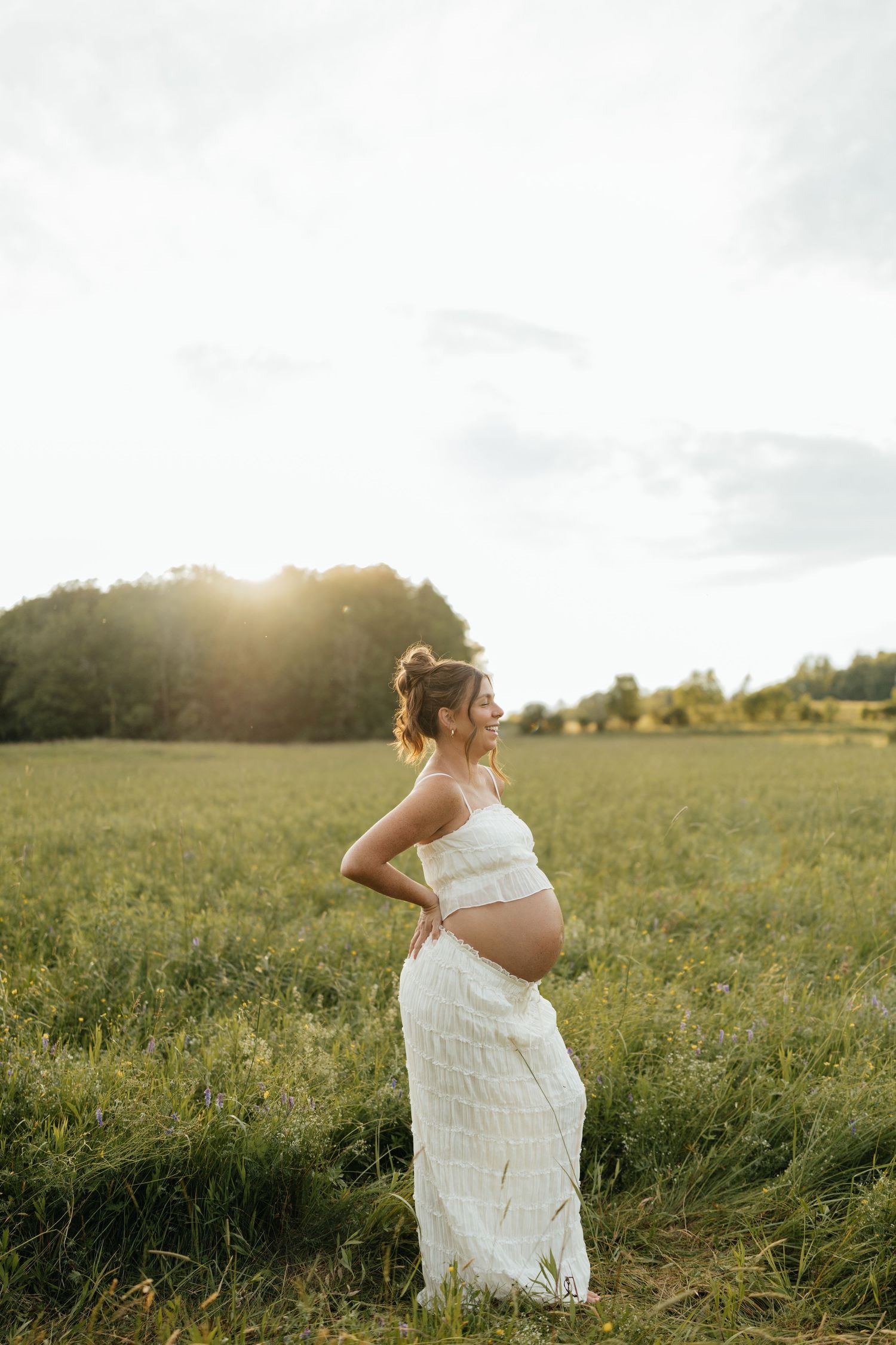 Pregnant woman in white dress standing in green field at golden sunset, cradling baby bump with hands on hips.