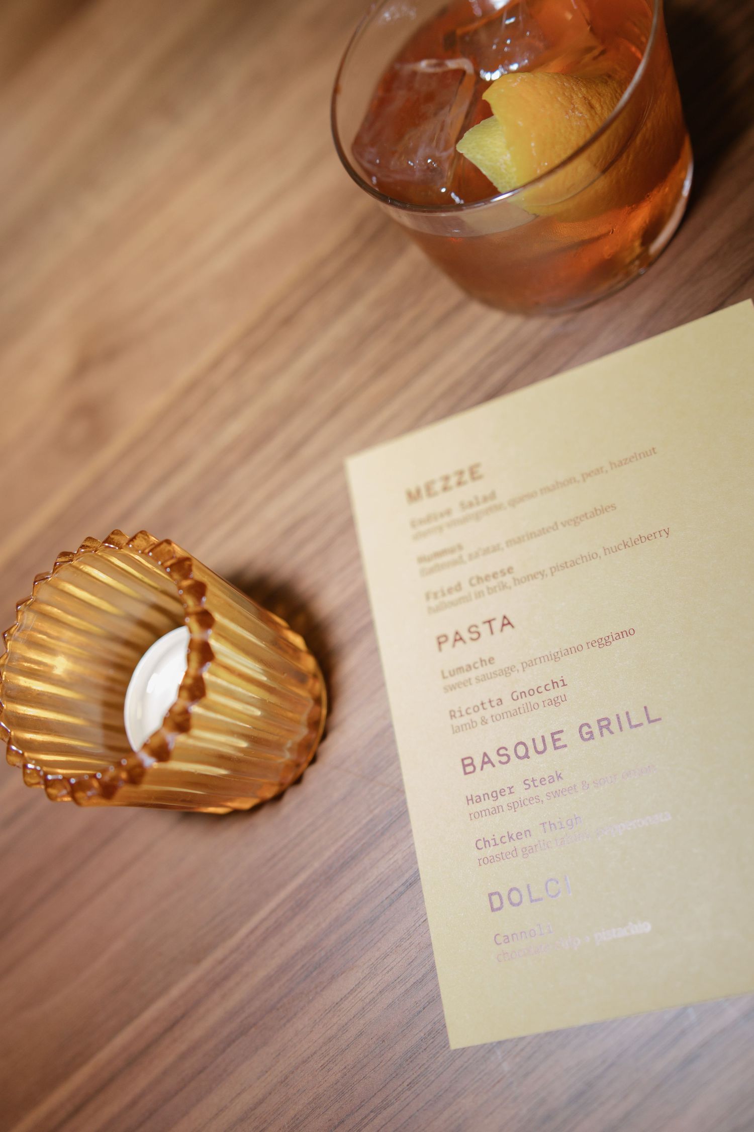 Restaurant menu beside a cocktail with ice and citrus garnish, and a gold scalloped dish on a wooden table.