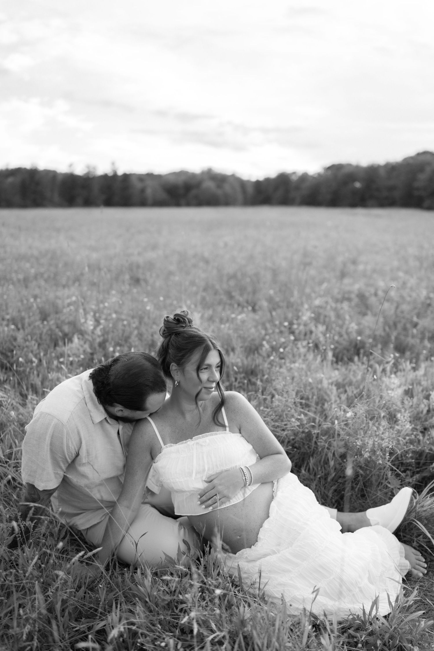 Black and white photo of a couple sitting in a field, woman in white dress cradling baby bump, man kissing her shoulder.