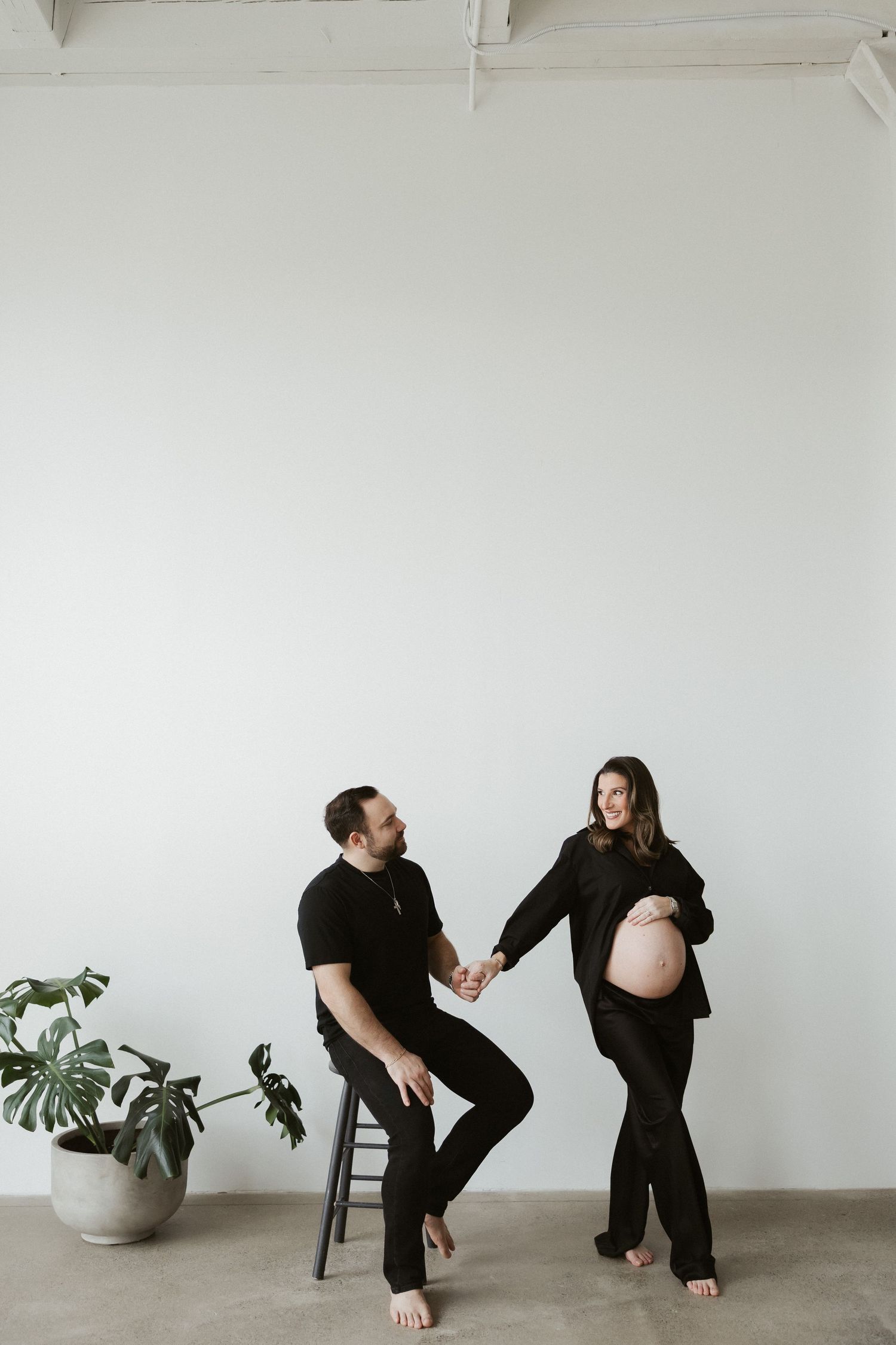 Expecting couple in matching black outfits holding hands during maternity shoot, woman displaying baby bump near monstera plant.