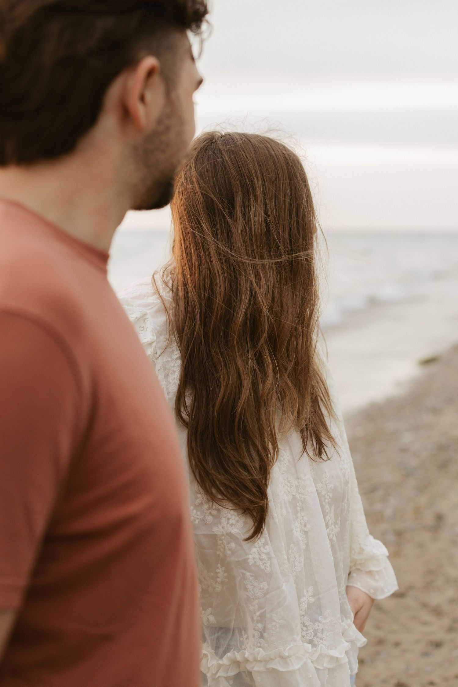 Couple standing together on a beach, woman in white lace dress with long brown hair, man in rust shirt, ocean view.