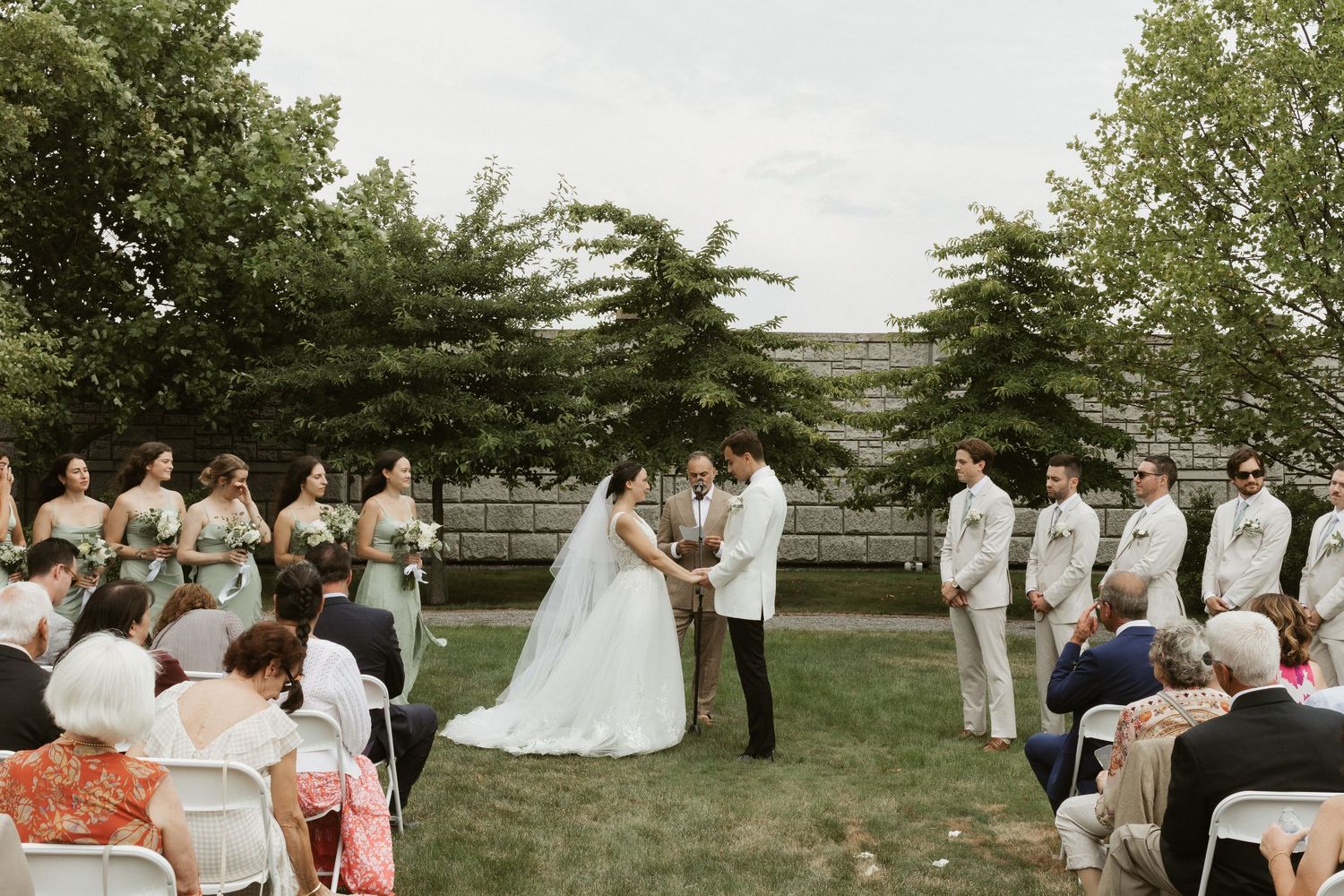 Wedding party gathers outdoors under trees for group photos during a summer celebration.