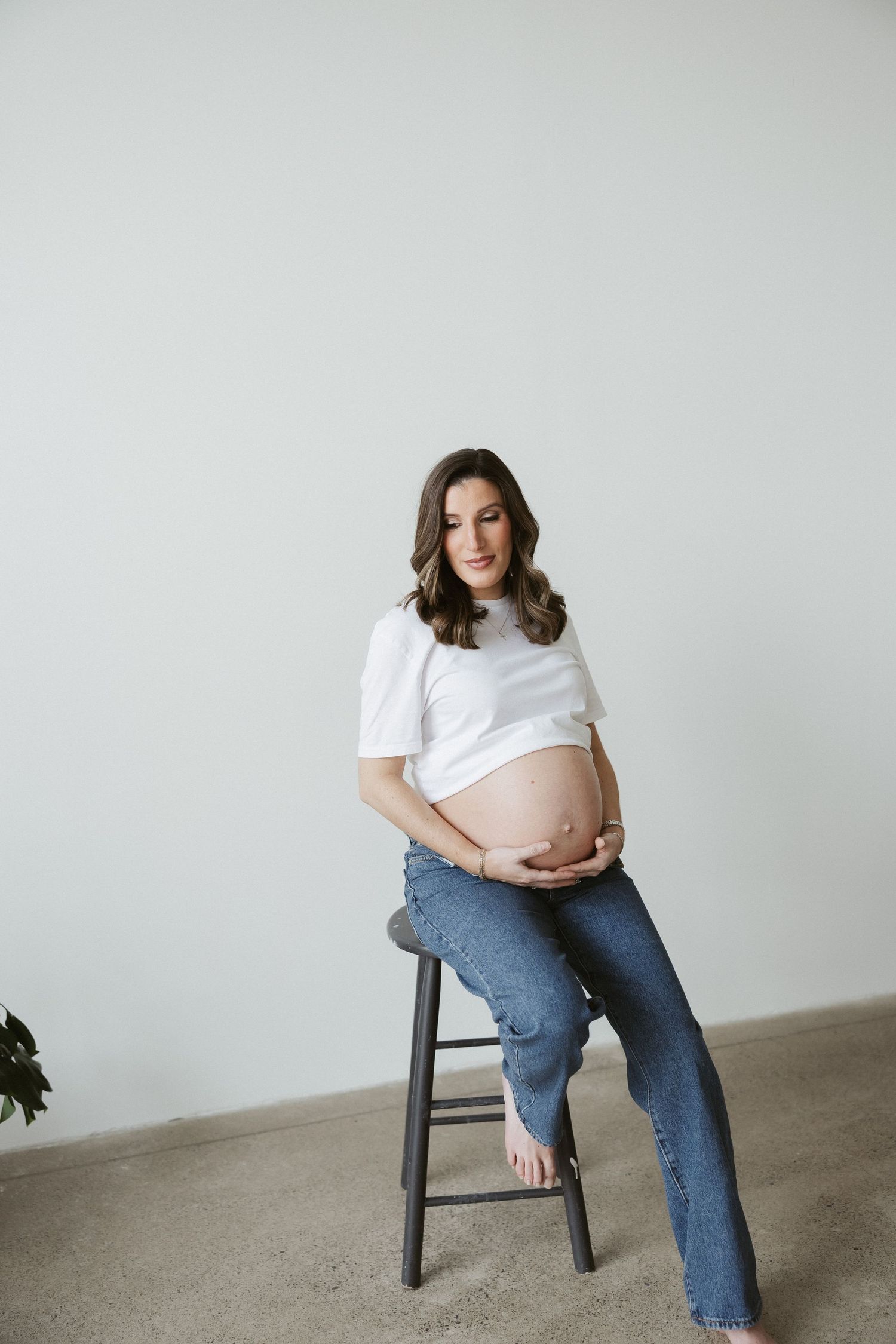 Pregnant woman in white crop top and jeans sitting on stool, cradling baby bump in a bright minimalist studio.