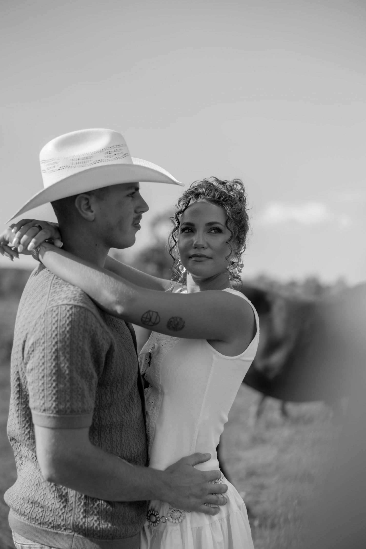 A romantic black and white photo of a couple embracing outdoors, with one wearing a cowboy hat and sweater vest.