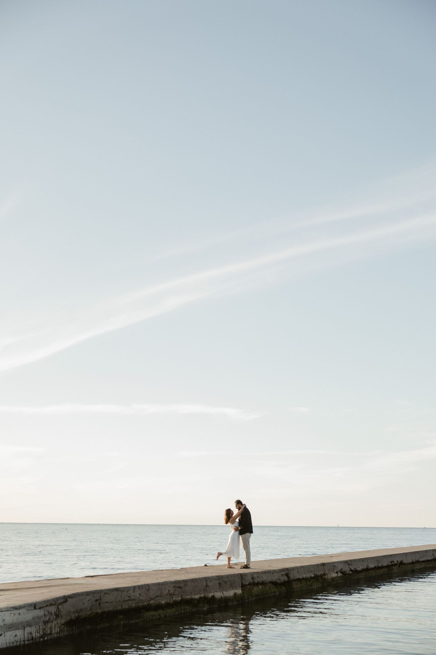 Silhouetted couple stands at the end of a long concrete pier extending into a calm ocean at sunset.