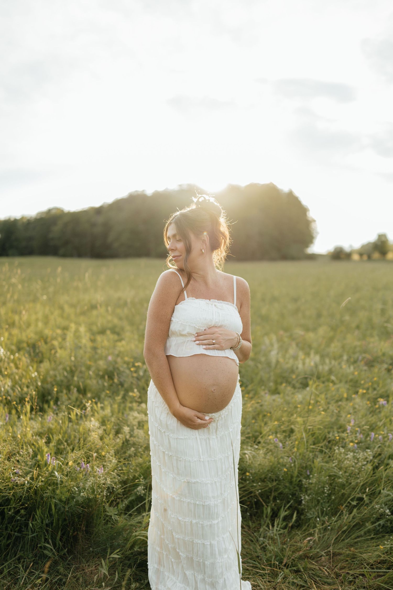 Pregnant woman in white dress holding baby bump in sunny green field.