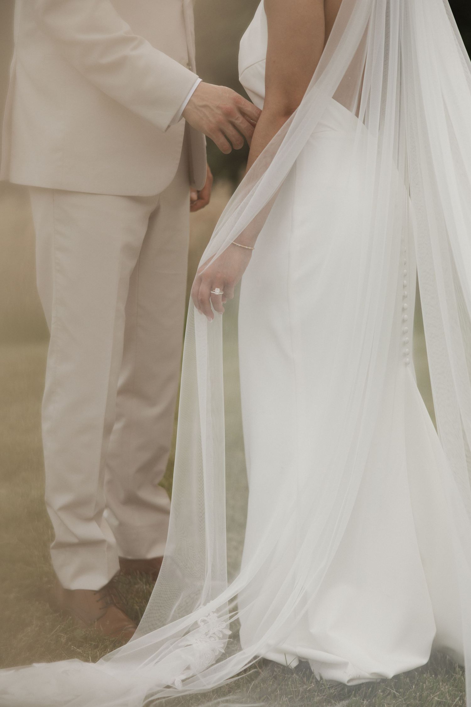 Bride in white gown and veil holds hands with groom in beige suit, both barefoot outdoors.