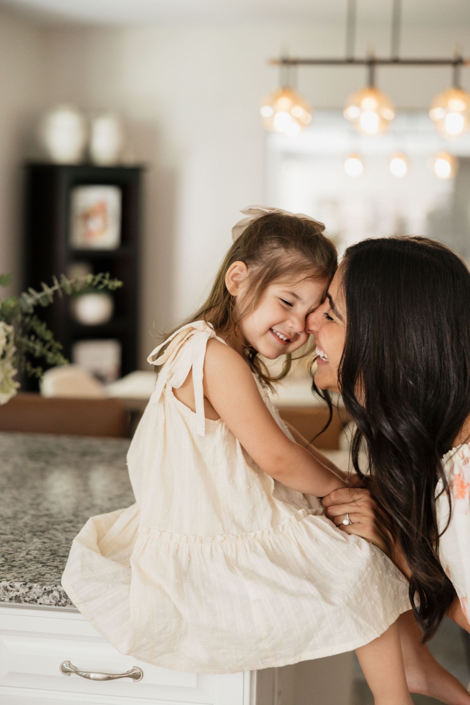 A mother and young daughter share a joyful moment in a bright kitchen, both wearing white dresses.