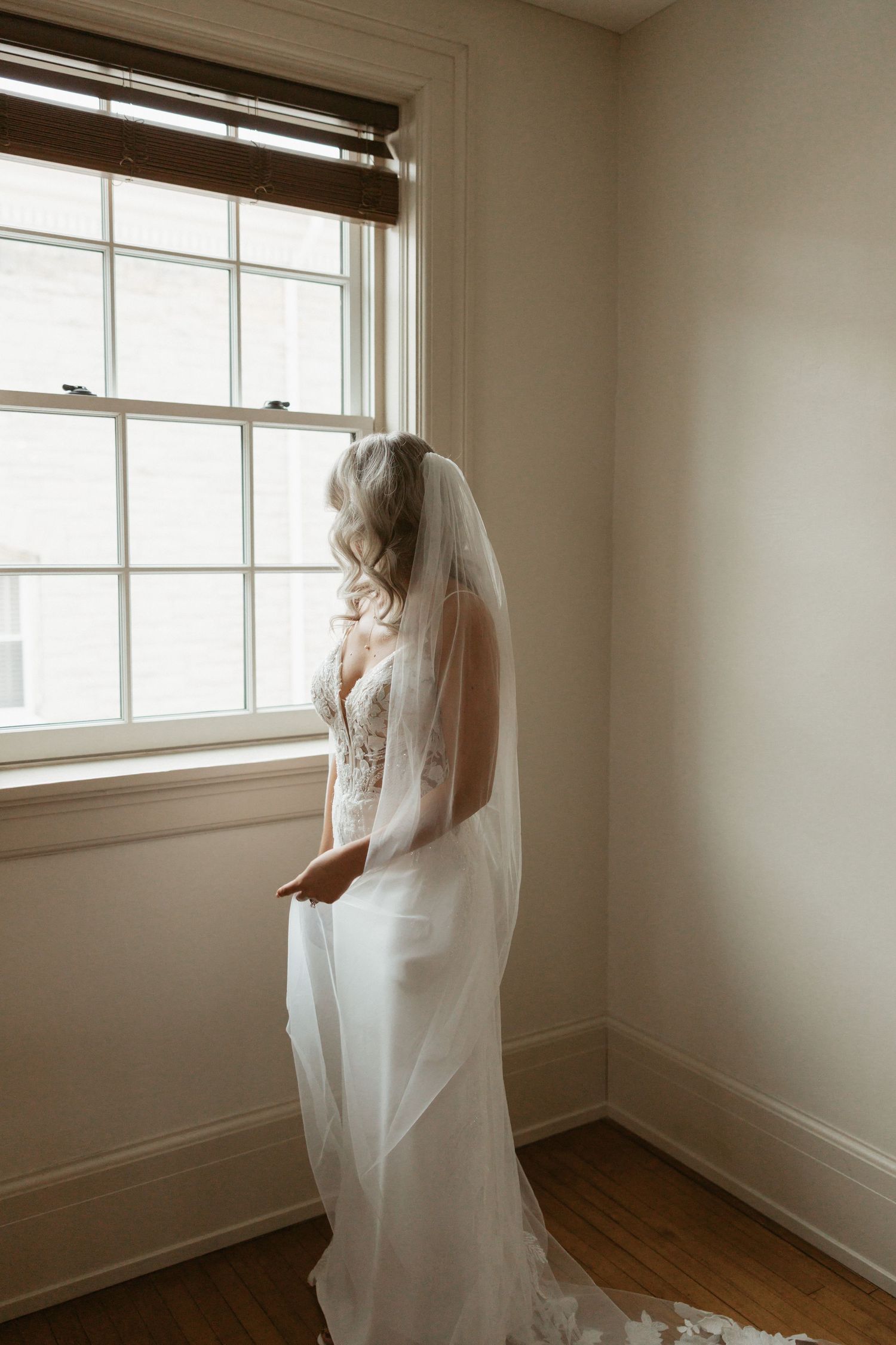 A bride in a flowing white veil stands near a window in an empty room with hardwood floors and white walls.