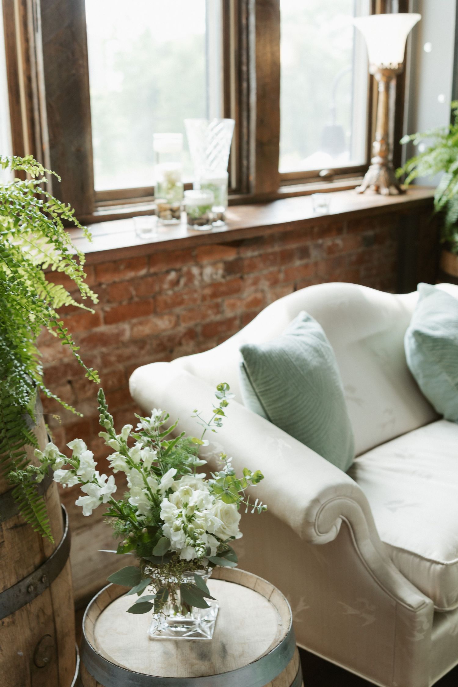 Cozy living room corner with white sofa, green pillows, and fresh floral arrangements by exposed brick windows.