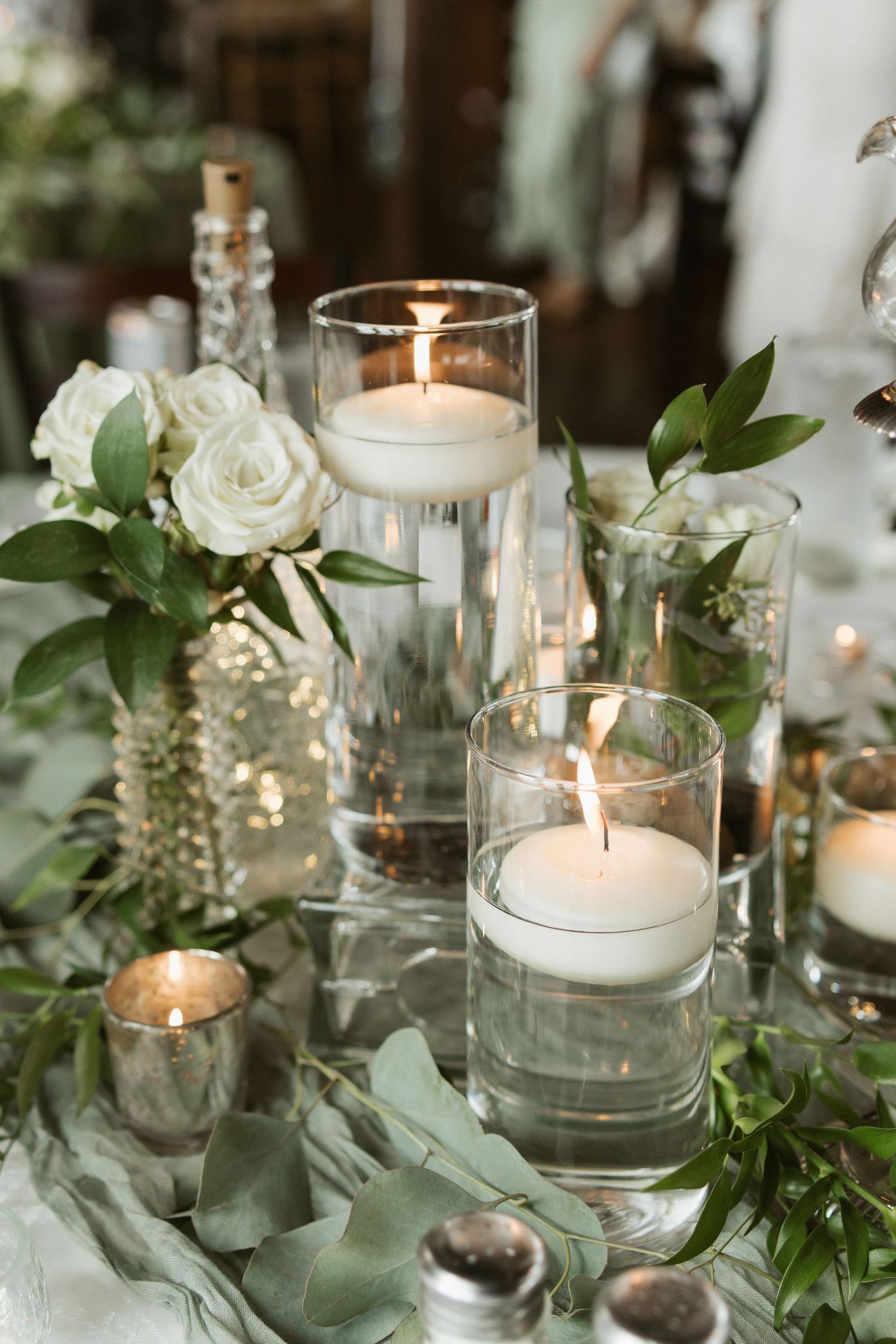 Elegant wedding table decor with glowing candles in glass holders surrounded by fresh greenery and white roses.
