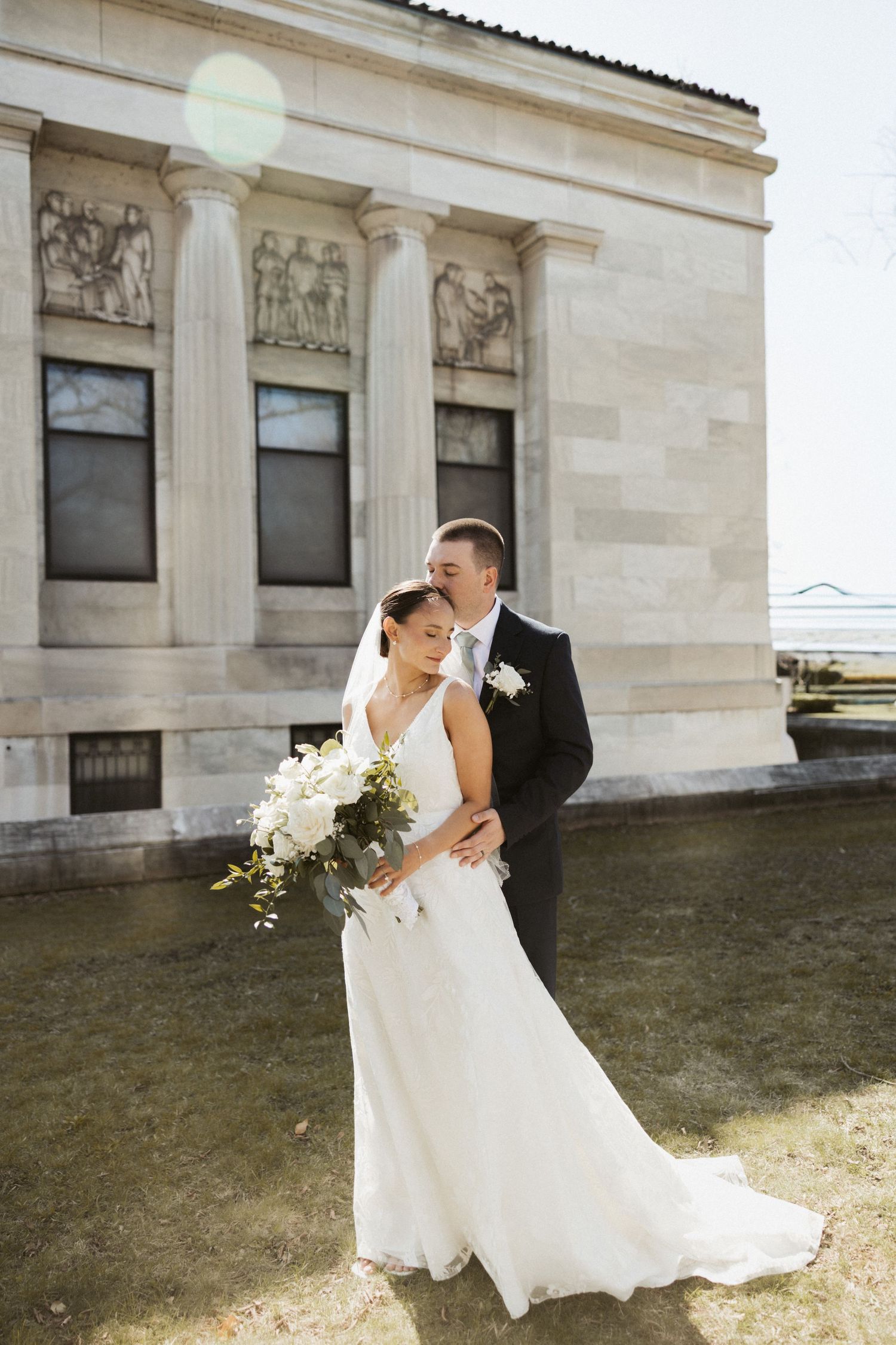 Bride and groom embrace outside a neoclassical building, bride holding white bouquet in flowing wedding gown.