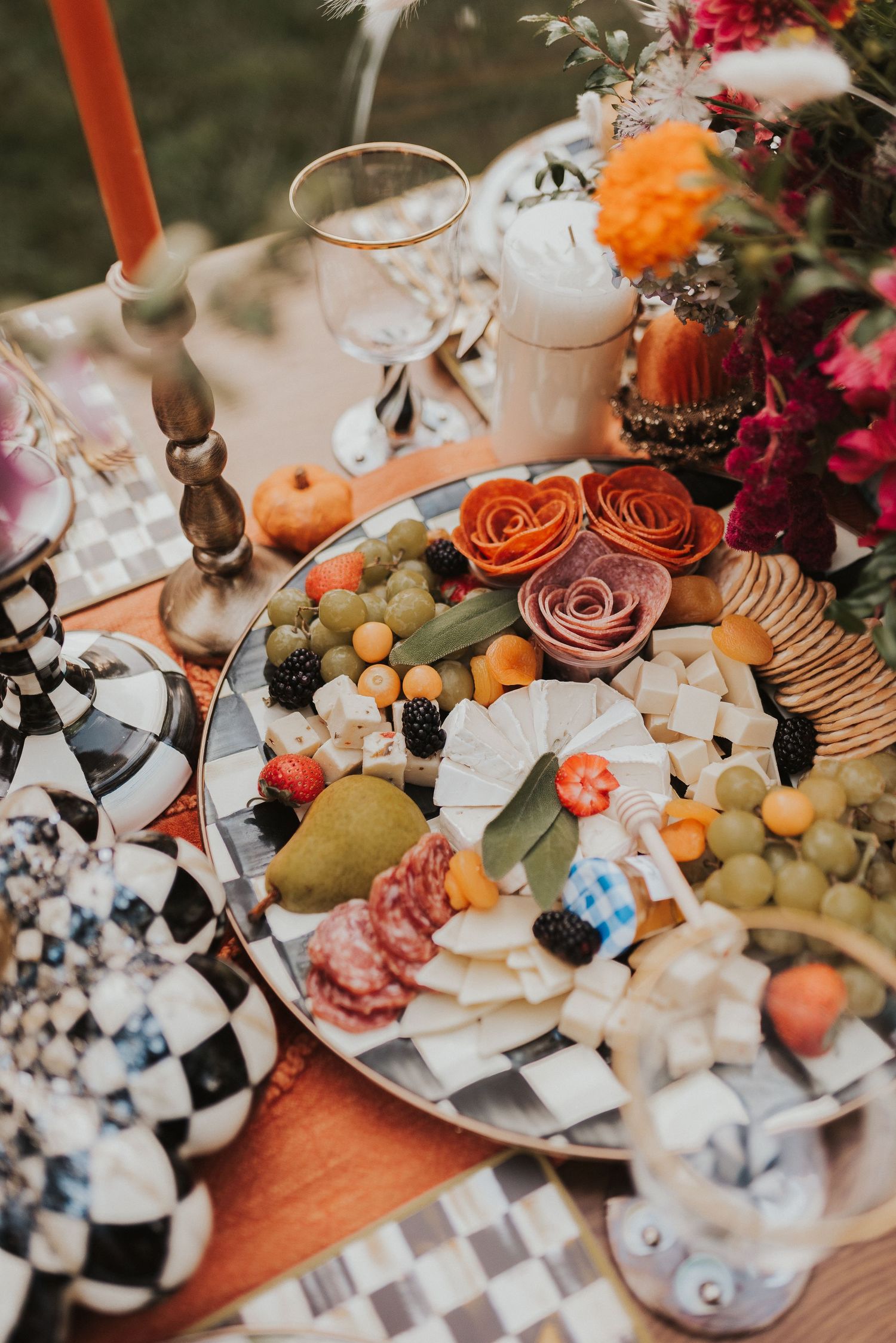 Elegant party spread featuring cheese boards, fresh fruits, roses, and decorative checkered elements on a wooden table.