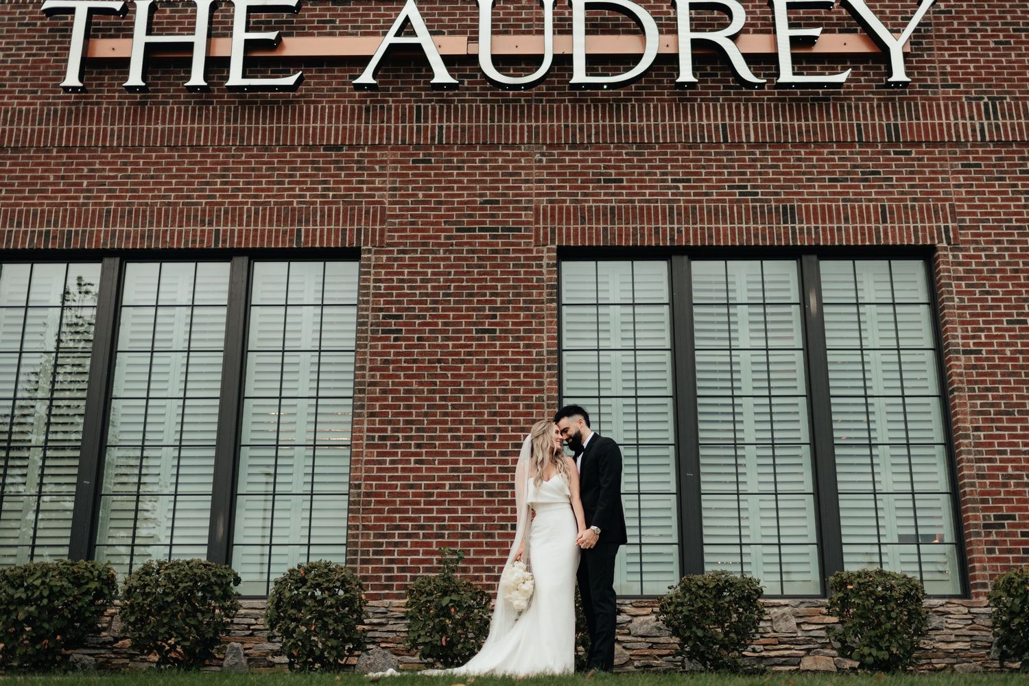 A bride and groom embrace in front of a brick building with 'THE AUDREY' signage and large industrial windows.