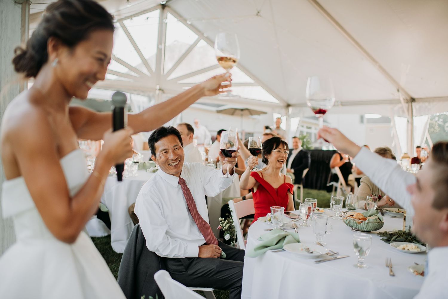 Wedding guests raise their glasses in a celebratory toast under a white tent during an outdoor reception.