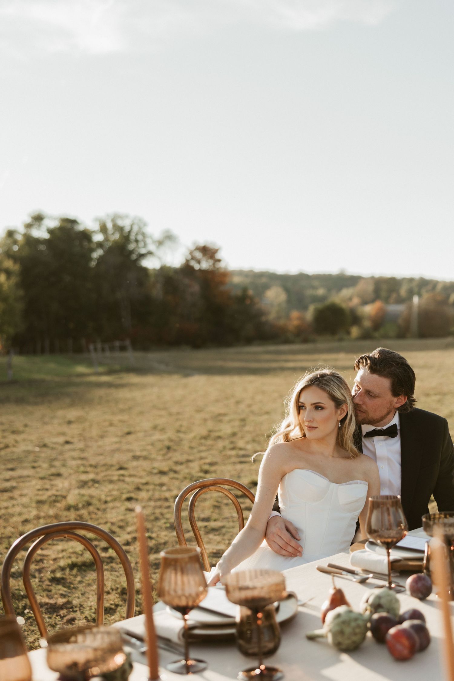 A rustic outdoor wedding reception table setup with wooden chairs and golden hour lighting in a pastoral field.