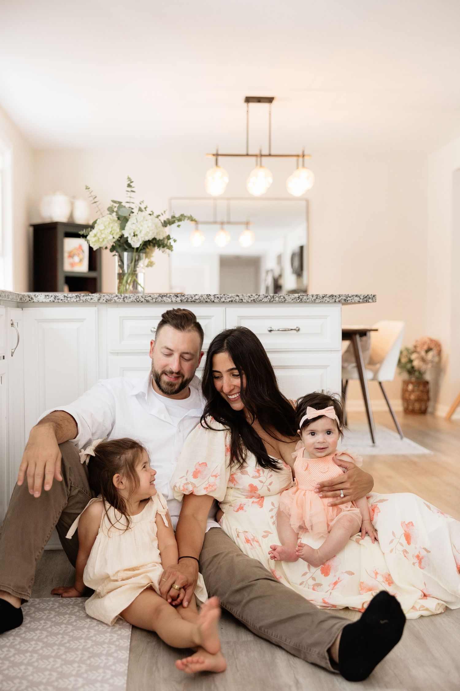 Happy family of four sitting on kitchen floor, parents smiling at baby and toddler daughter in bright modern home.