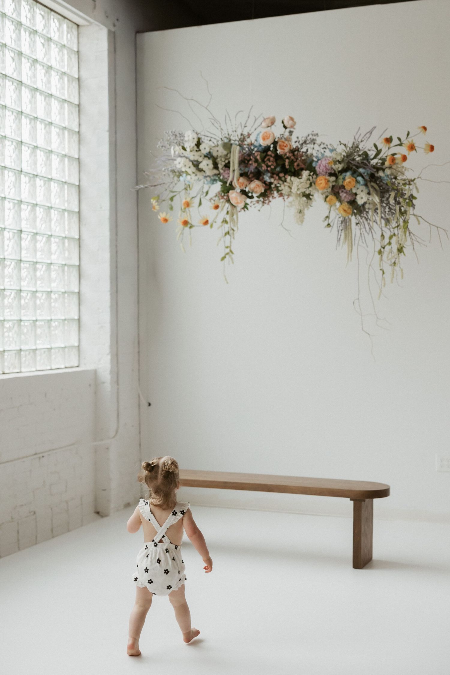 A toddler gazes at a stunning floral wall installation above a wooden bench in a bright white studio.