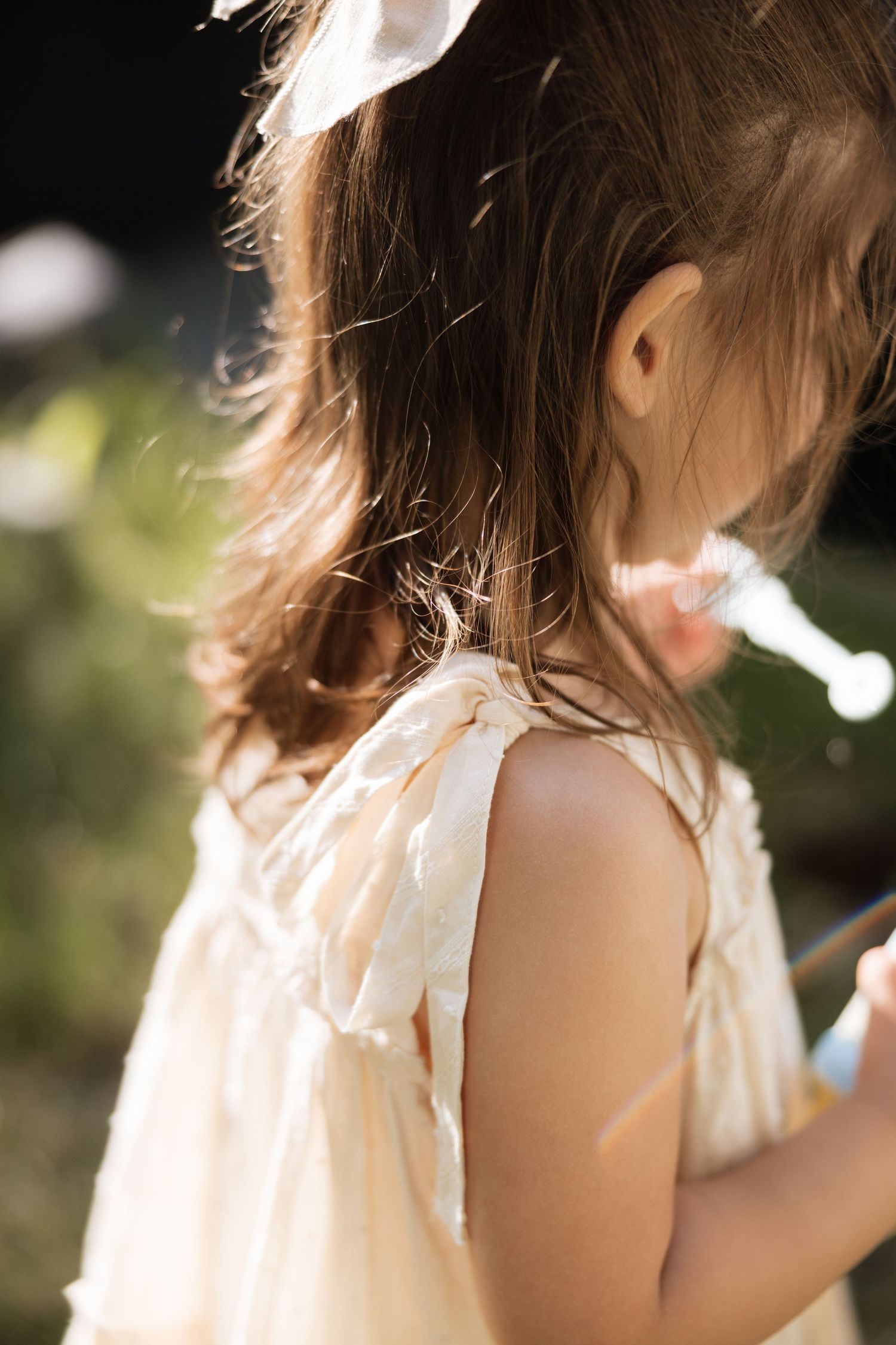 Young girl in cream dress with white bow hair accessory stands outdoors in warm sunlight.