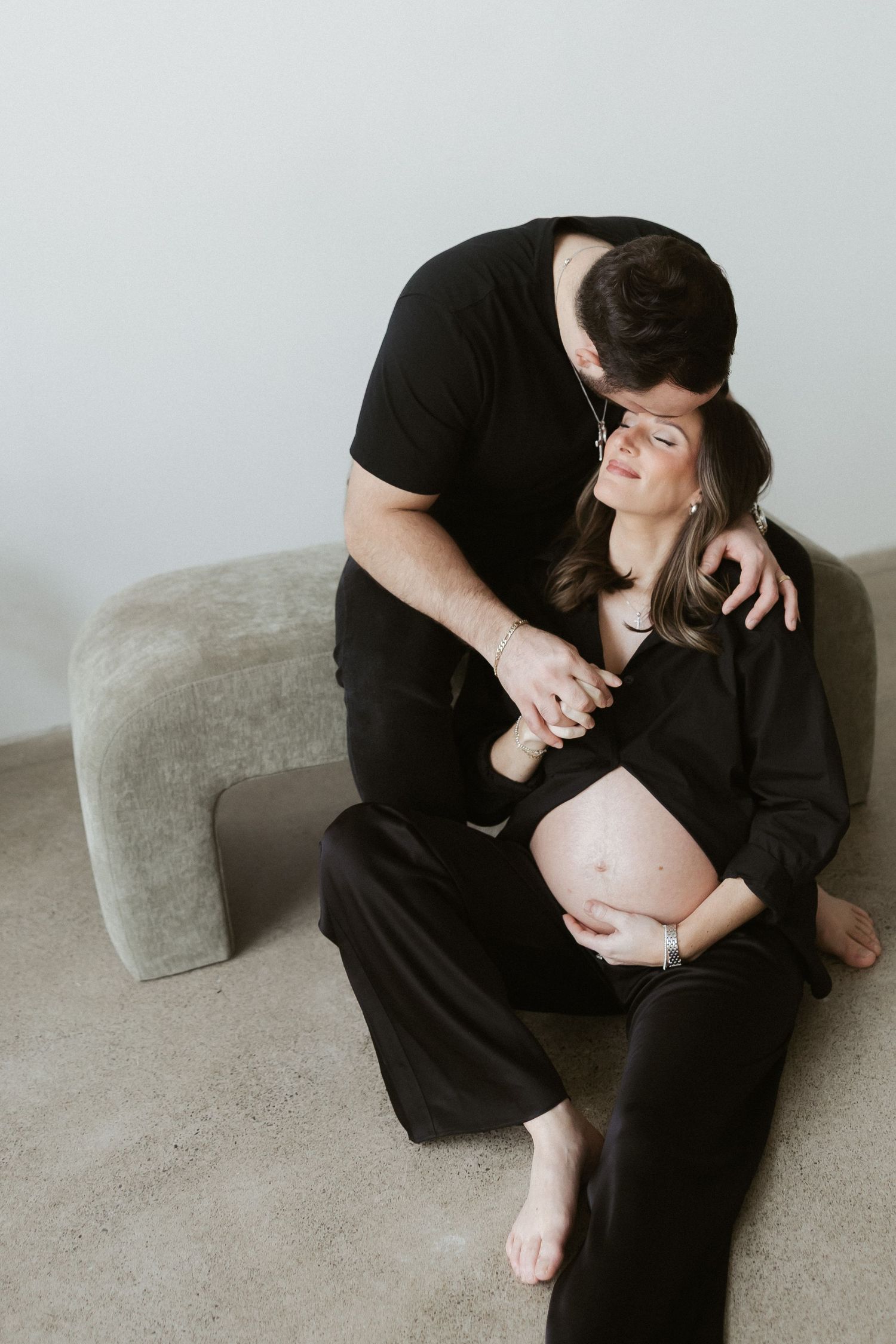 A couple in black outfits shares a tender moment, the man kissing his pregnant partner's head while holding her bump.