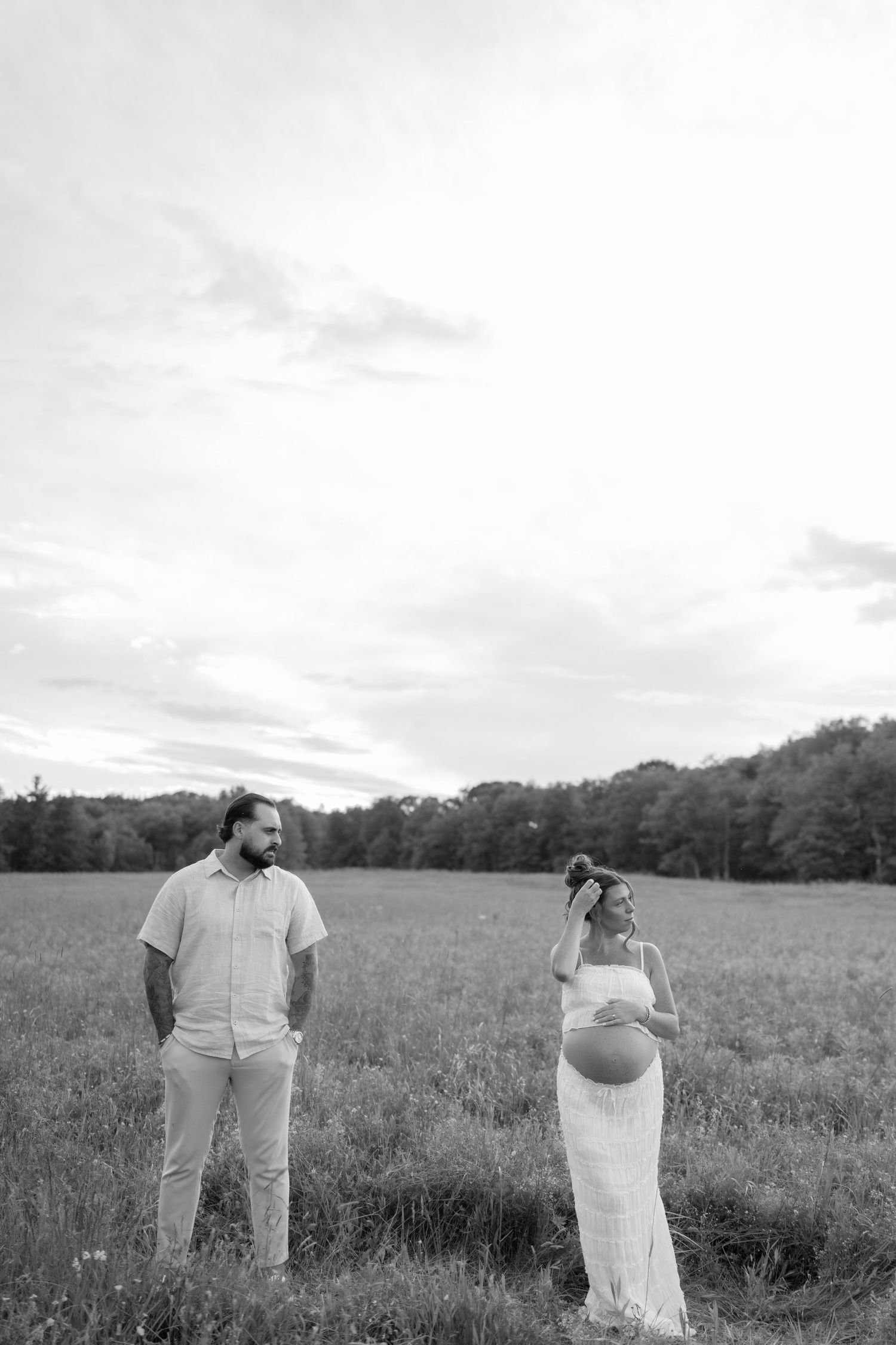 Black and white maternity photo of pregnant woman and bearded man standing in open field with trees in background.