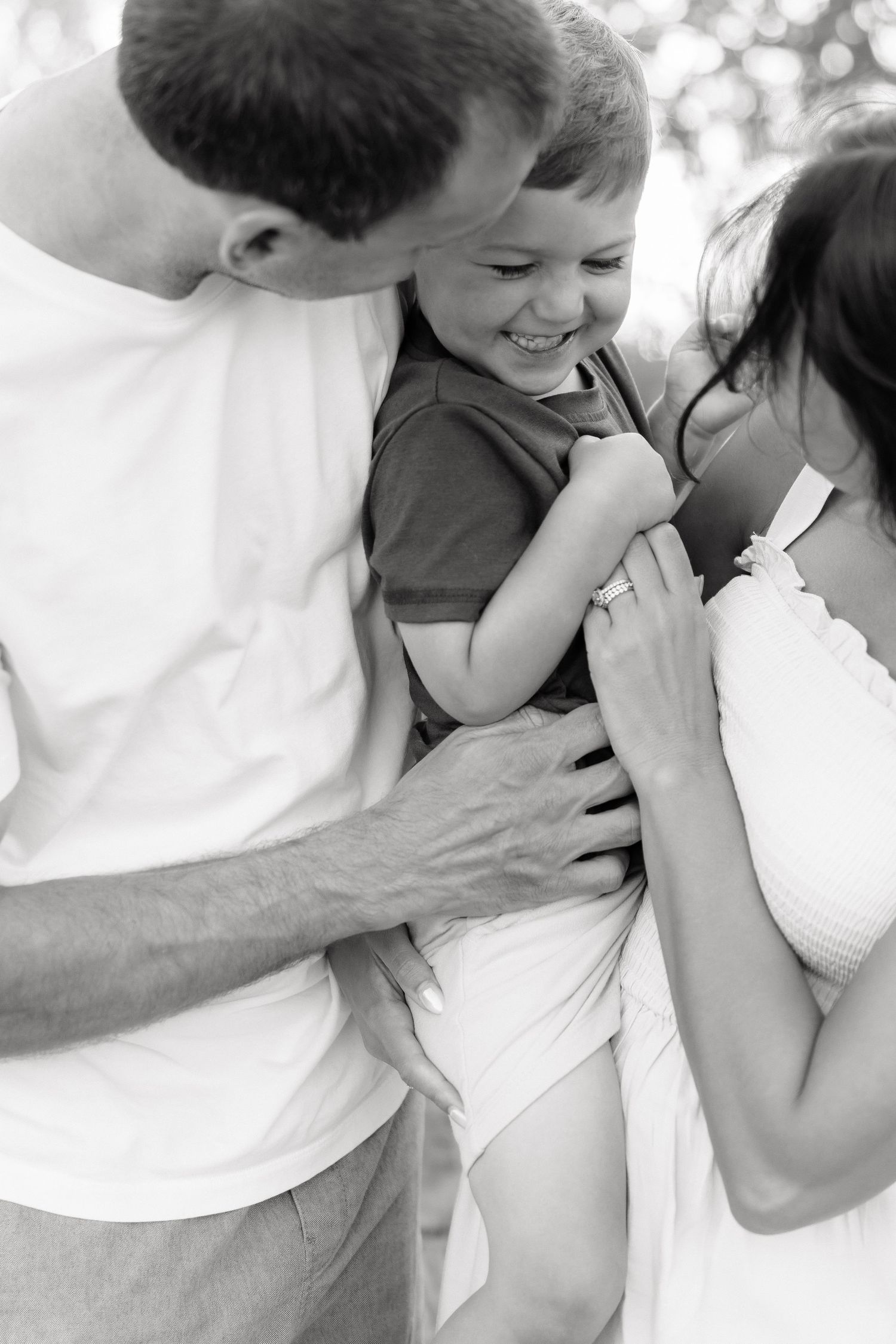 Black and white photo of happy family, parents holding laughing toddler boy outdoors in warm embrace.