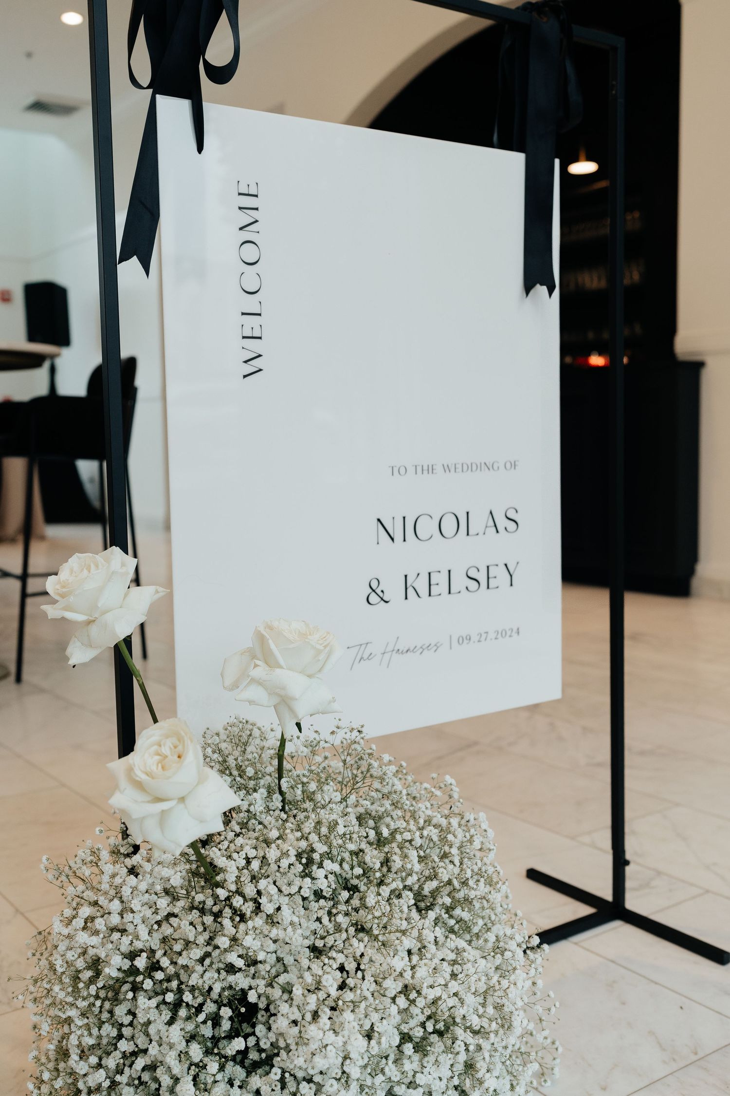 Elegant wedding signage displayed with white roses and baby's breath floral arrangements on stands against a white wall.