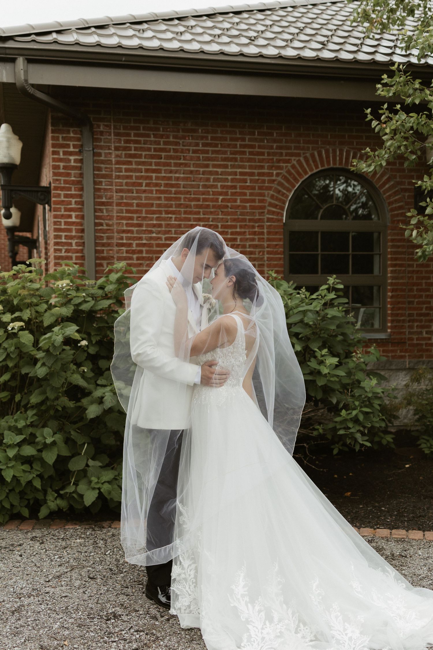 A romantic wedding moment captured outside a brick building with a flowing white veil and elegant dress.