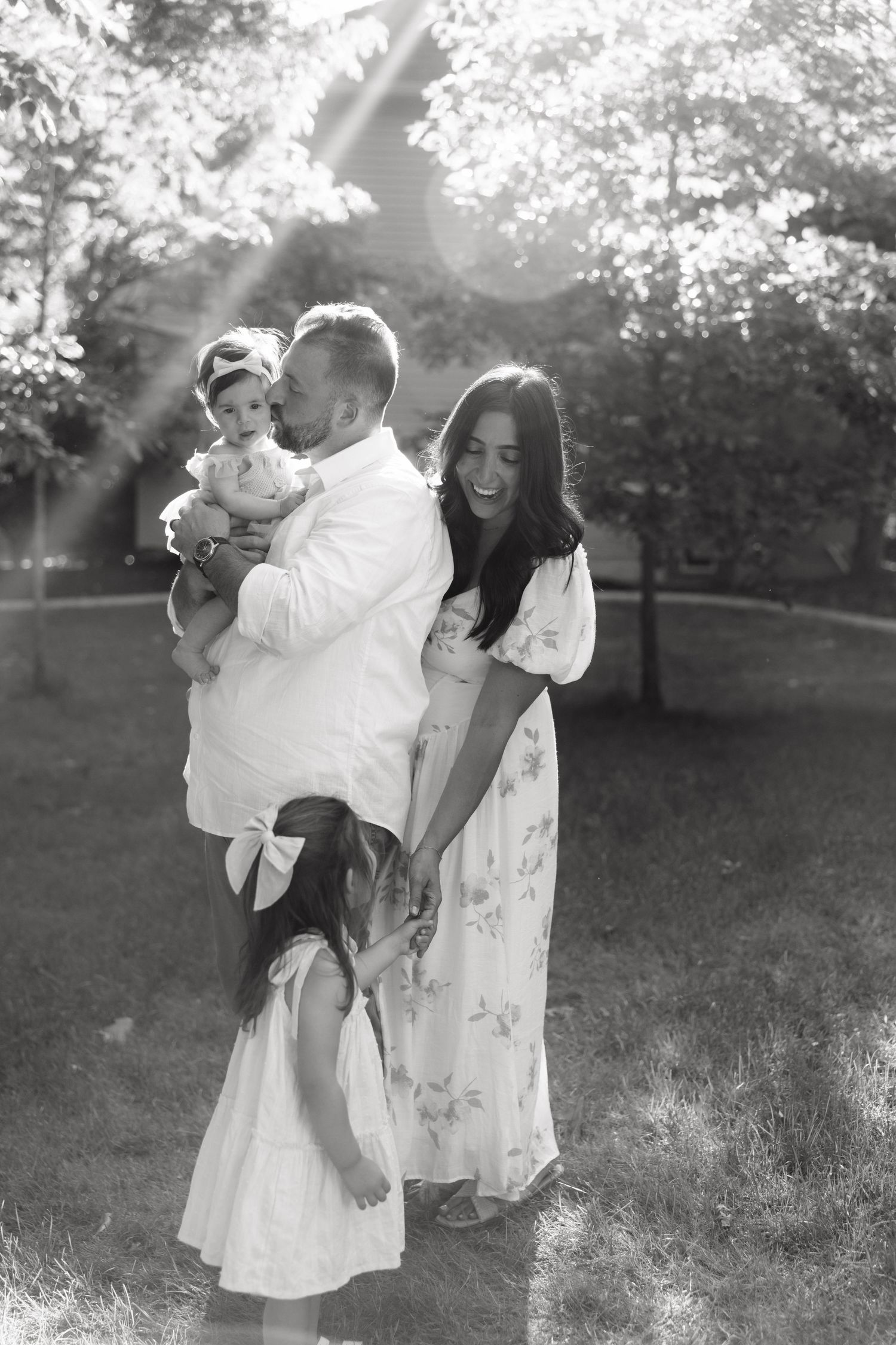 Black and white family photo of parents with two young daughters in a sunny park, laughing and holding hands.