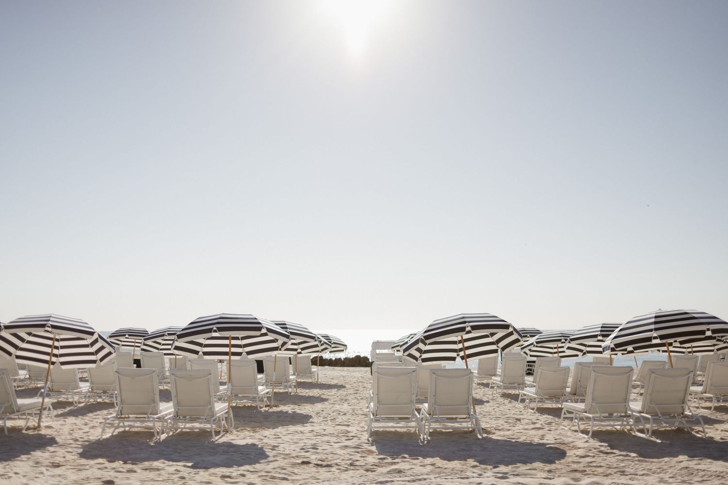 Rows of striped beach umbrellas and white lounge chairs on a sunny, sandy beach under a bright, hazy sky.