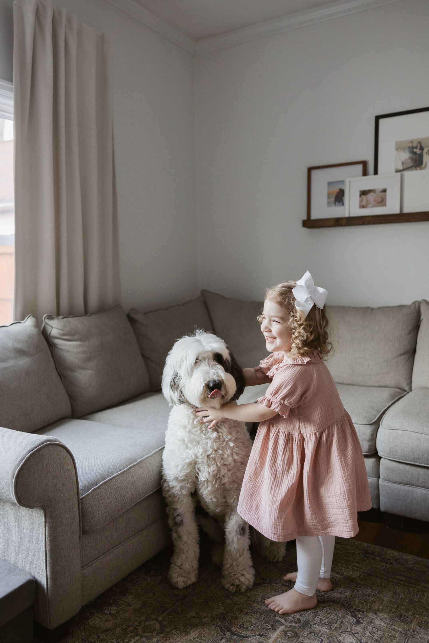 A young girl in a pink dress with a white bow hugs a large white fluffy dog in a cozy living room.