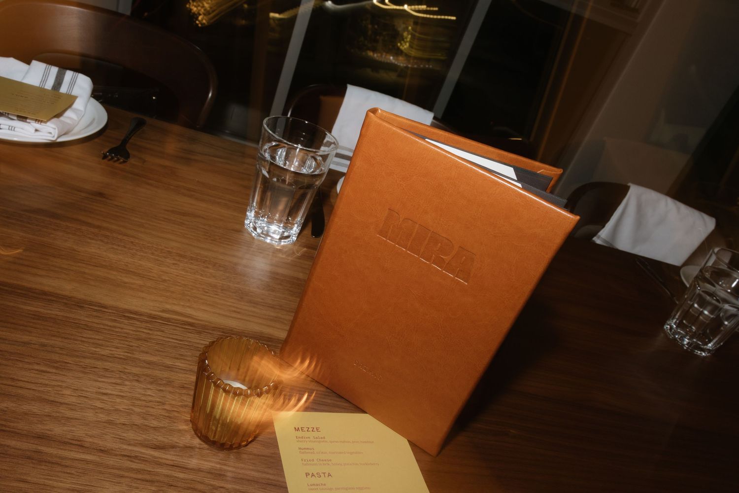 Restaurant table setting with leather menu, glass of water, and candle holder on wooden table.