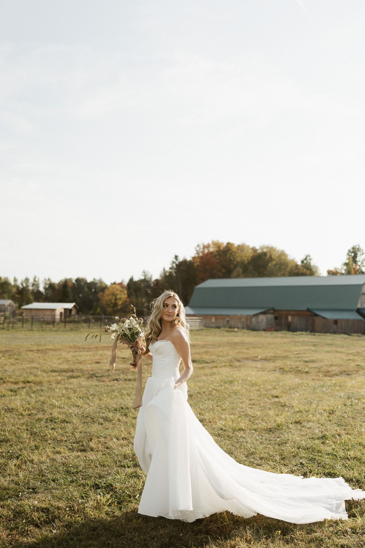 A bride in a flowing white wedding dress twirls gracefully in a golden sunlit field with farm buildings in the distance.