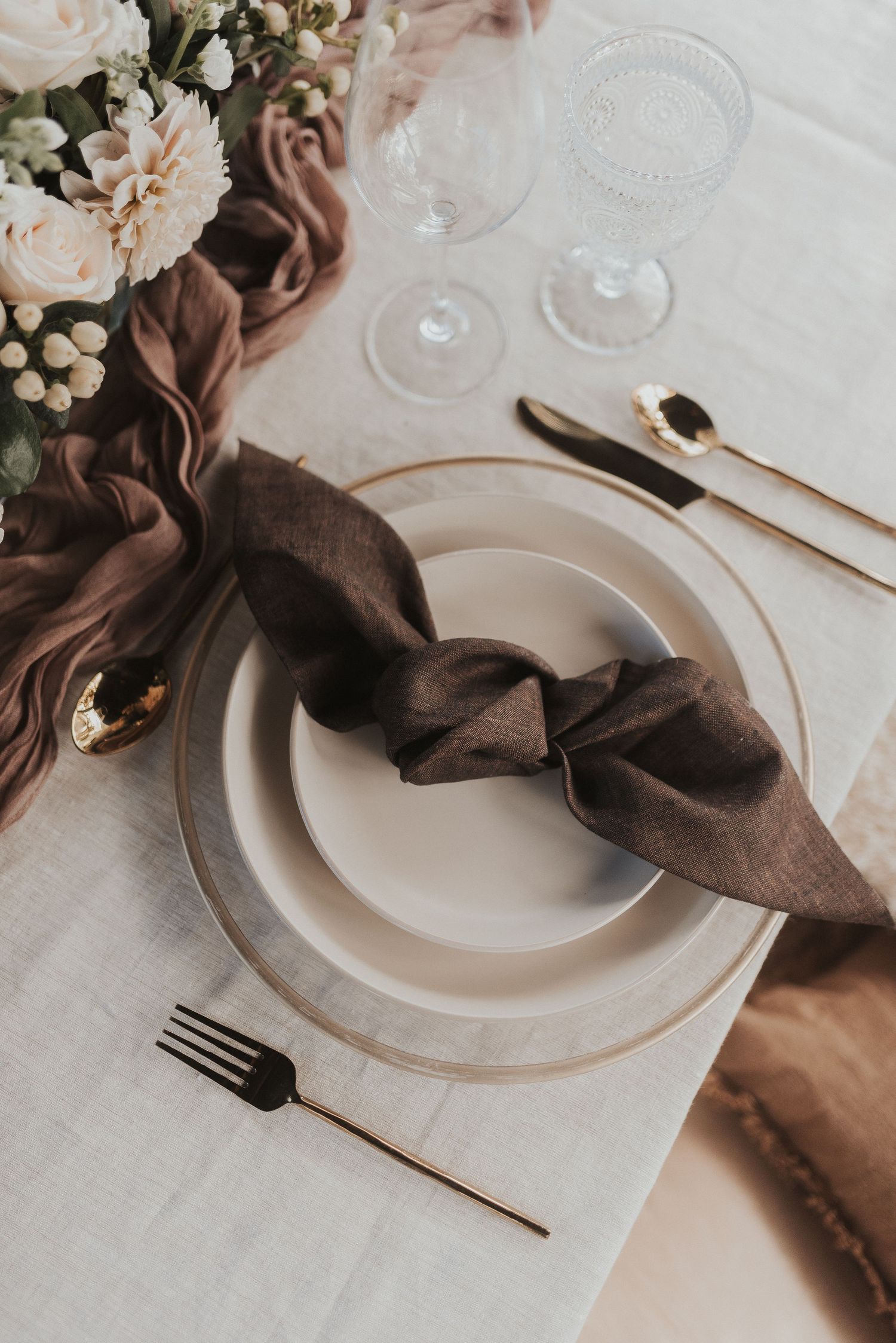 Elegant table setting with brown linen napkin, white plates, and soft pink floral accents on neutral tablecloth.