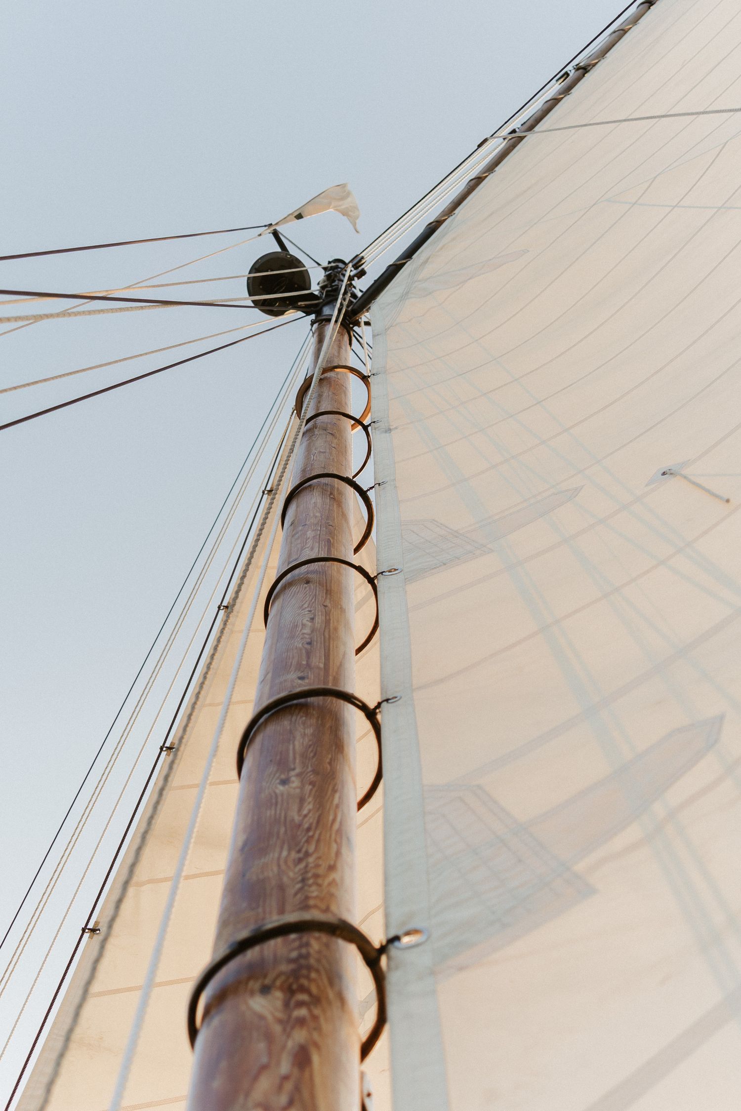 Close-up view of a wooden sailboat mast with white sail and rigging against a bright sky.