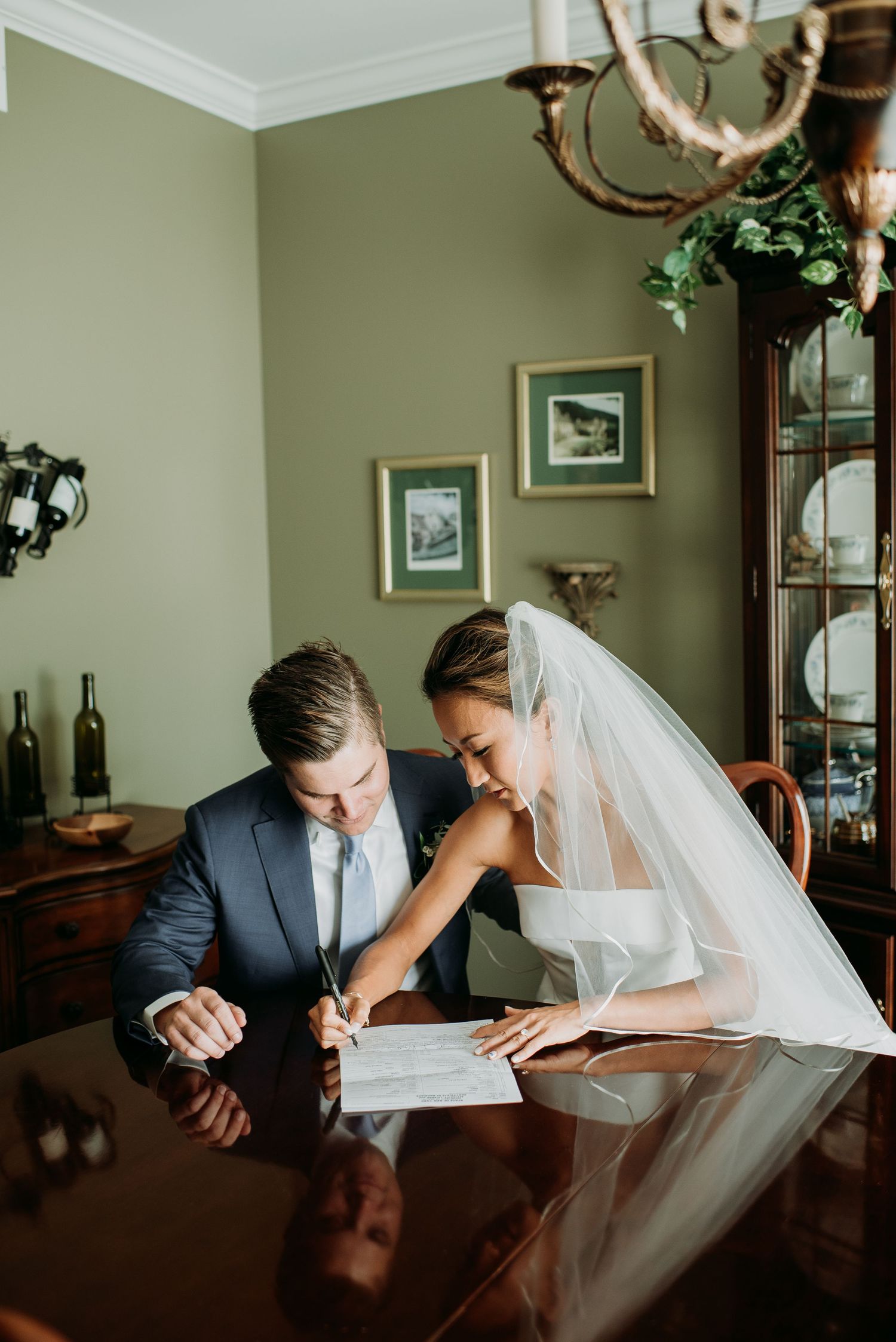 A couple in wedding attire shares an intimate moment while signing documents at a wooden table.