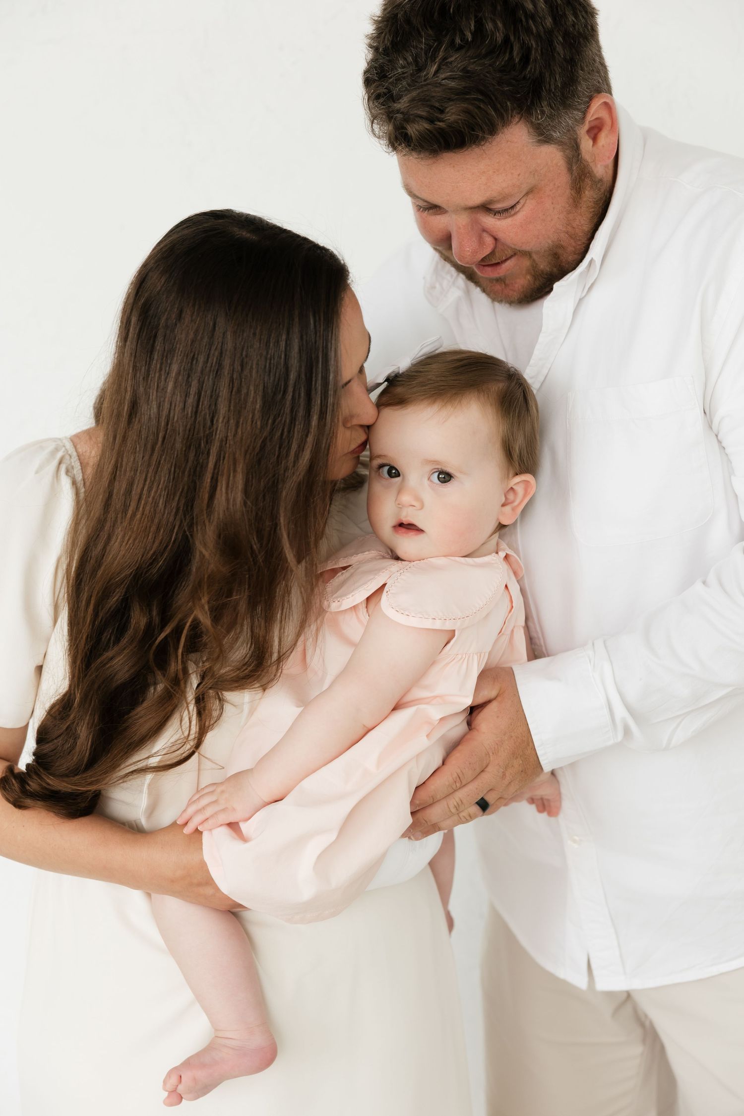 A couple in white clothing lovingly holds their baby girl in a pink dress against a white background.
