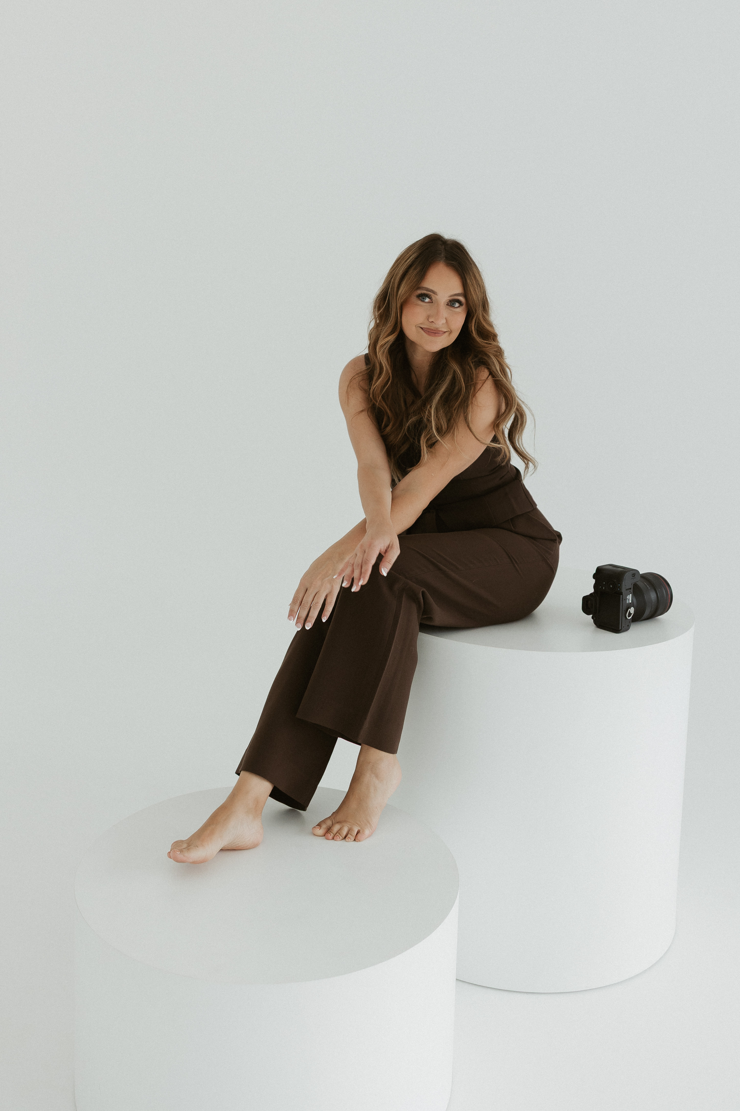 A woman in a brown jumpsuit poses gracefully on a white curved platform in a minimalist studio setting.
